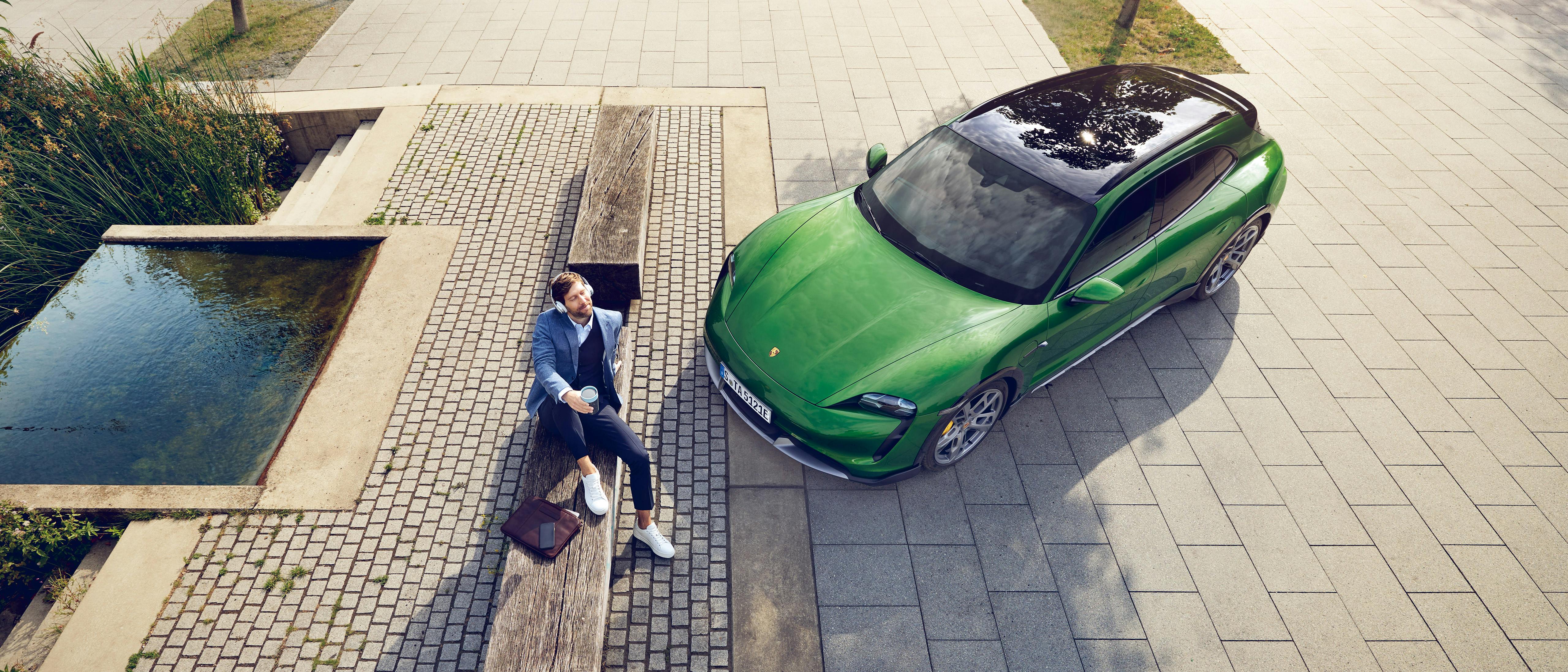 A man with a coffee-to-go mug sits on a bench next to a fountain, a green Porsche parked next to him.