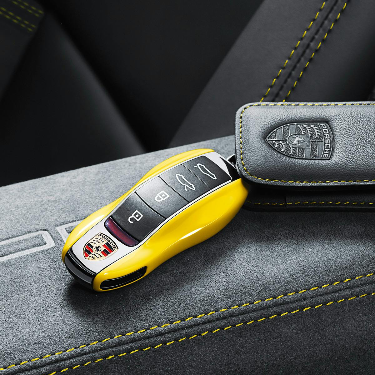 Yellow Porsche car key fob with logo and buttons placed on a gray interior surface with yellow stitching, accompanied by a leather key holder.