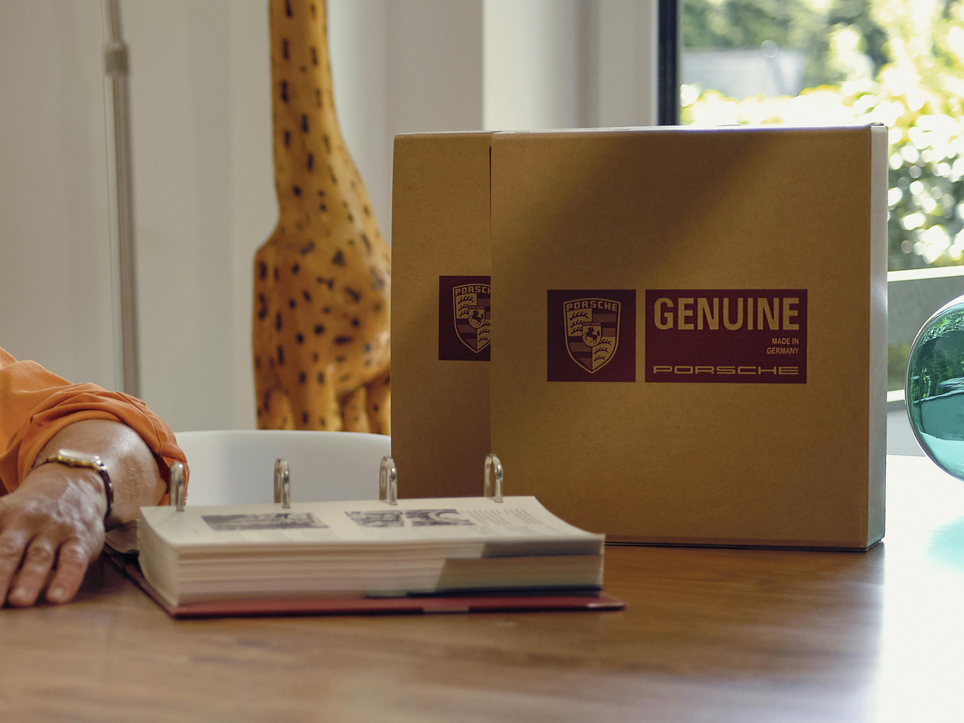 A folder and two packages labelled with the Porsche logo and the word GENUINE lie on a desk.