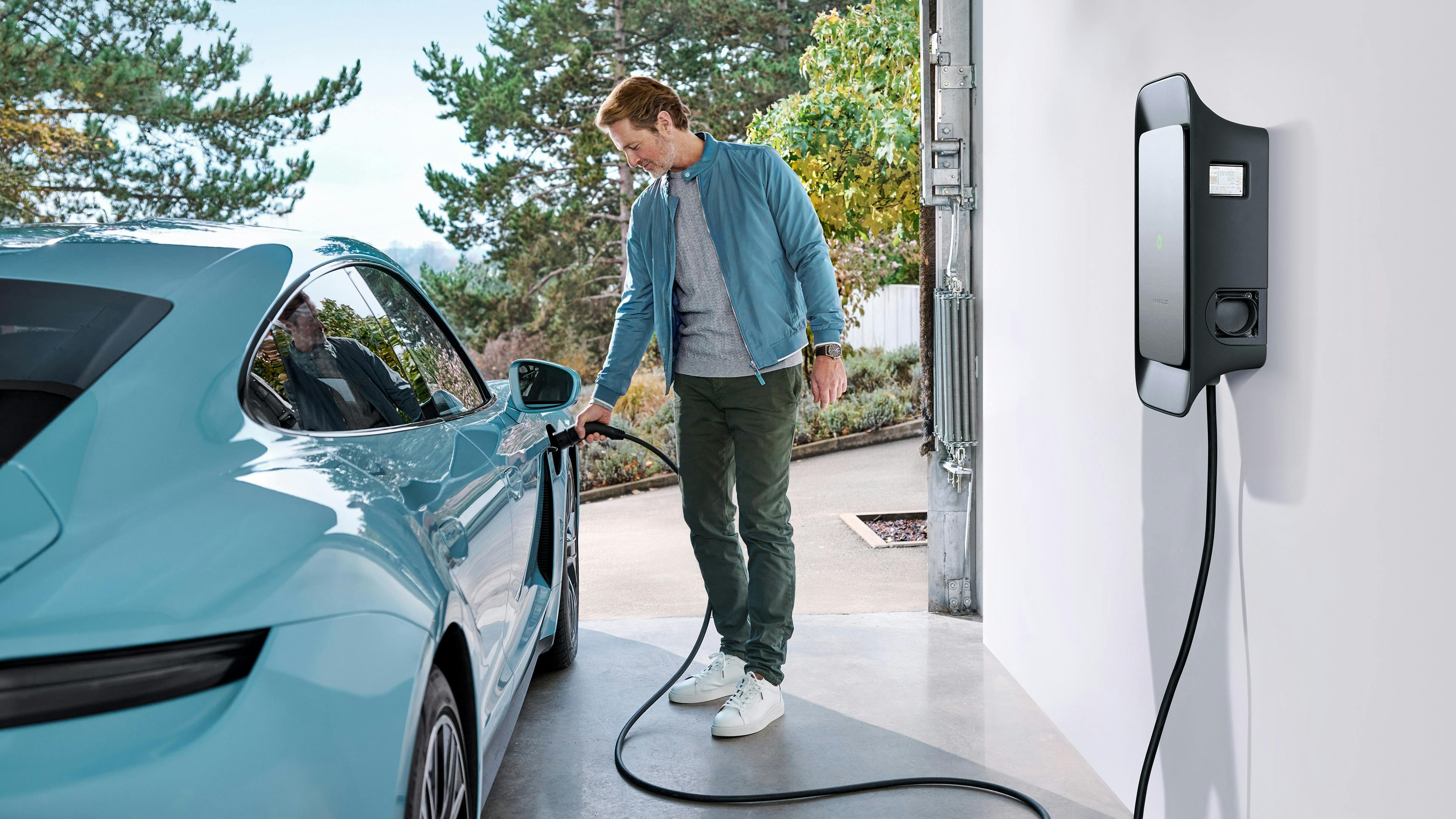 Man in blue shirt next to blue Porsche Taycan charging in garage.