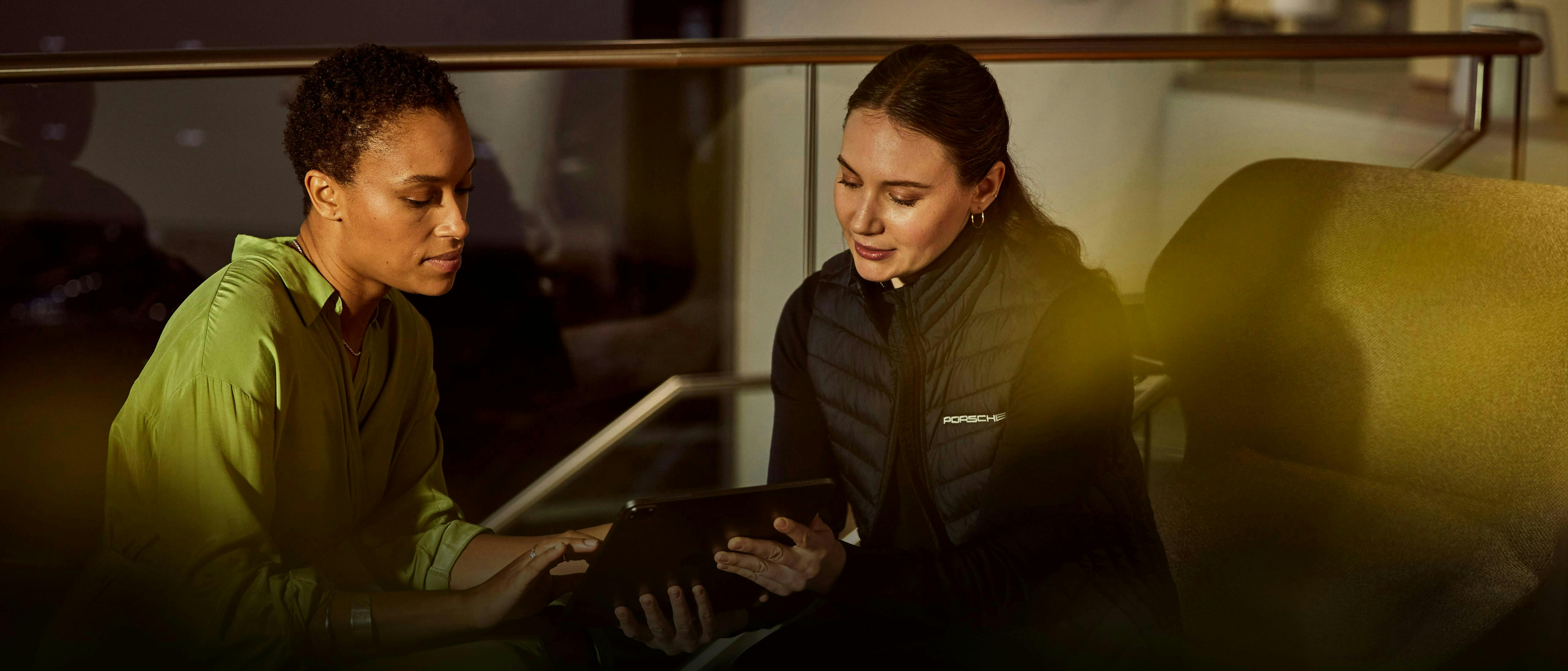 Two women sit in modern lounge collaborating on tablet, woman on right wears Porsche vest with logo.