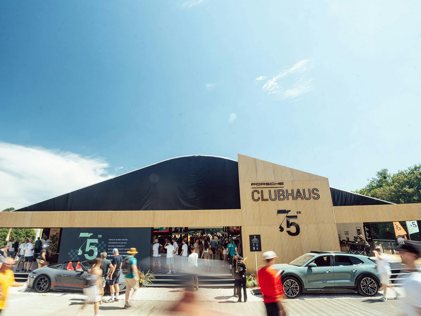Porsche Clubhaus exterior at Goodwood Festival of Speed, marking 75 years of Porsche in the UK, with visitors and a Porsche parked outside.