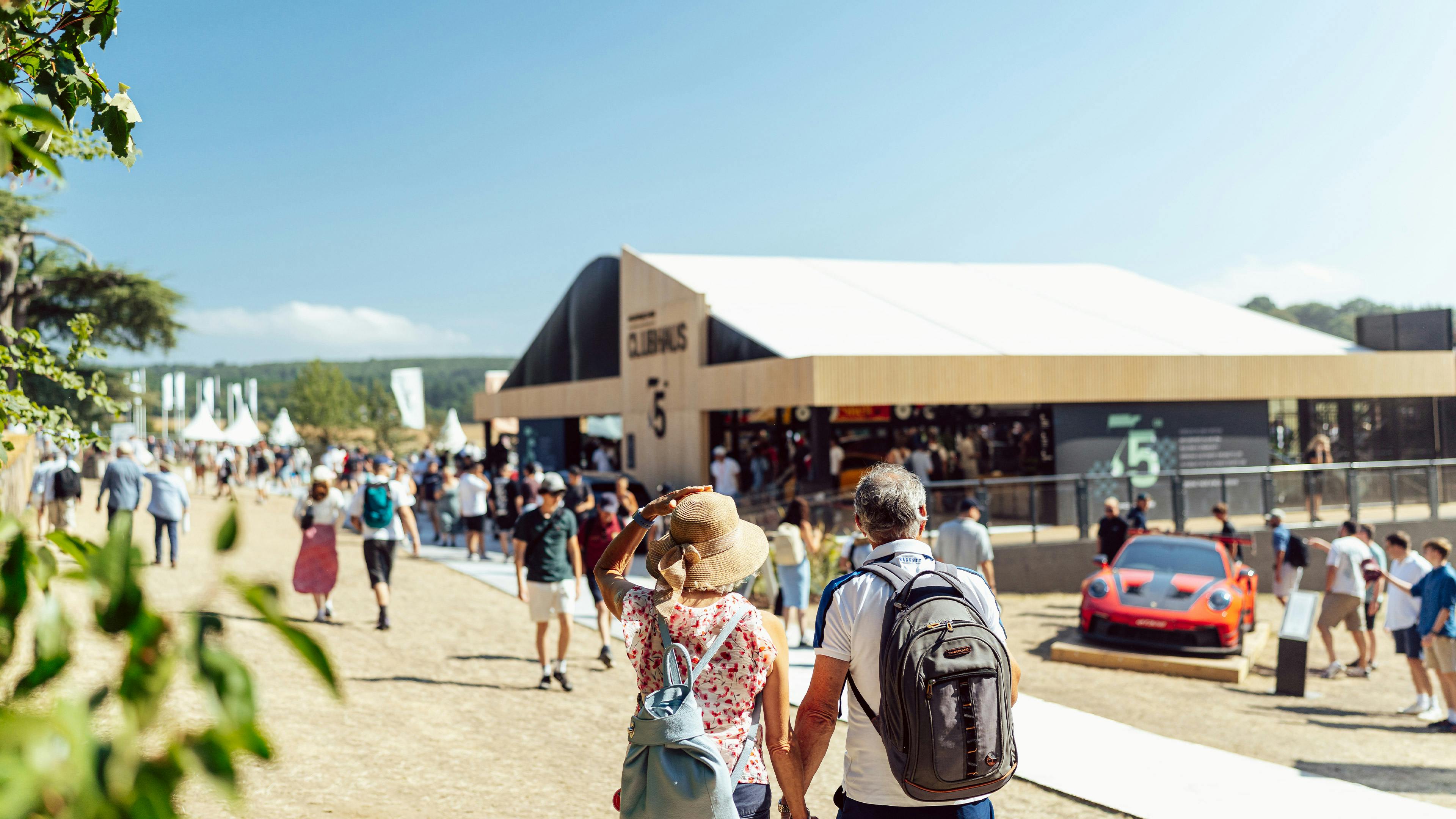 Festival-goers walking towards the Porsche Clubhaus on a sunny day at Goodwood, with a red sports car in the background.