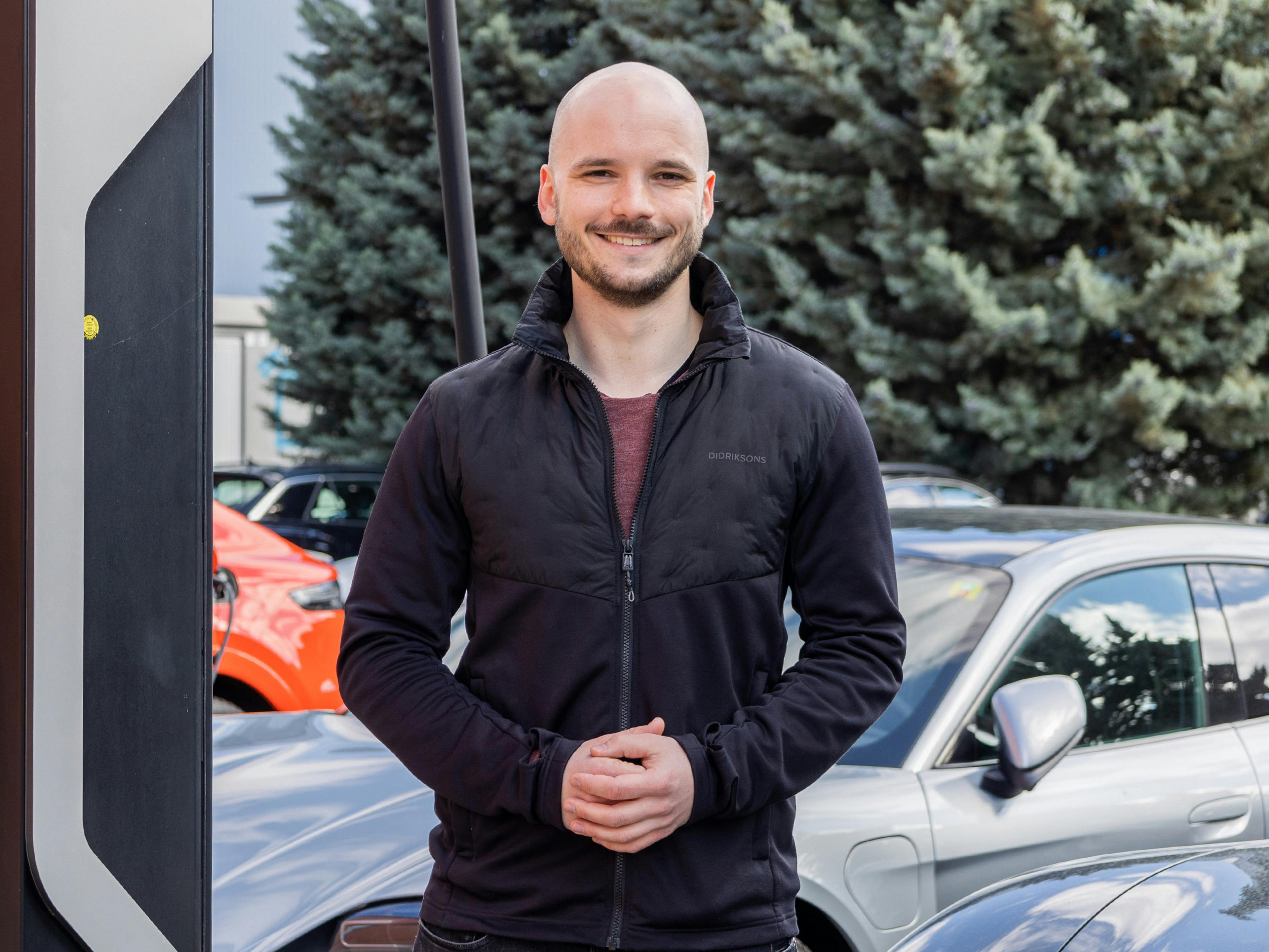 Der Porsche Mitarbeiter Tobias steht neben einer Ladesäule und lächelt in die Kamera.