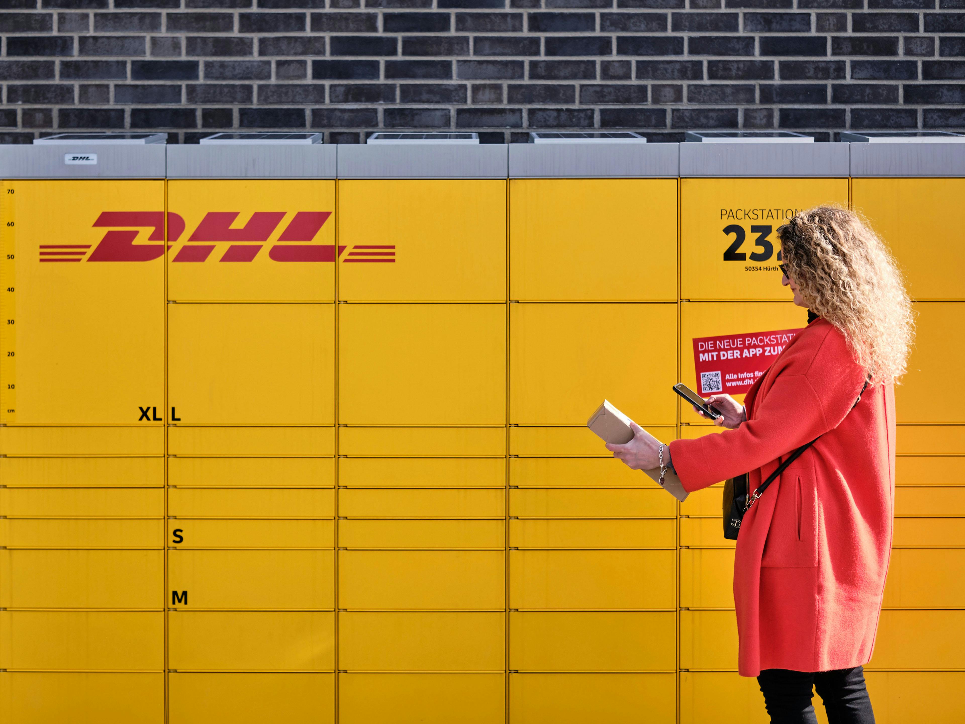 Blonde Frau in roter Jacke vor gelber DHL Packstation.