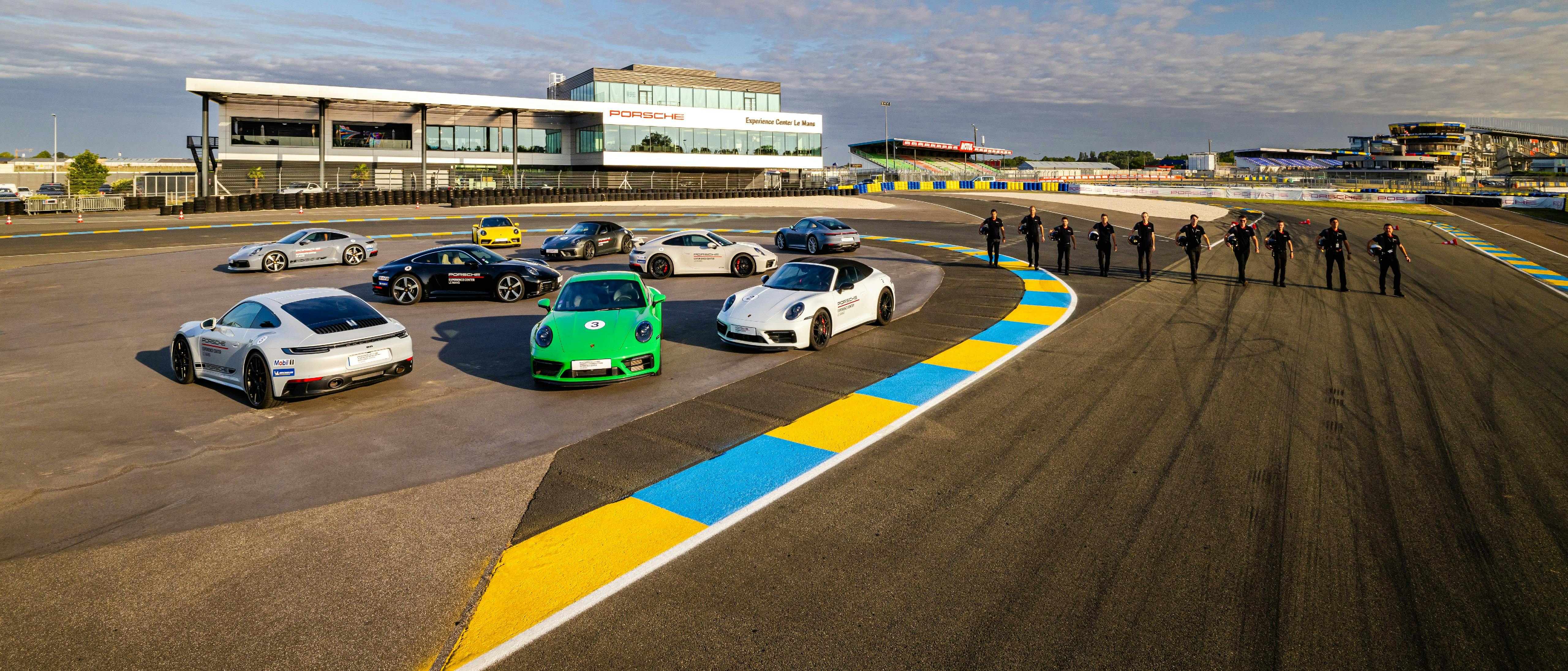 Porsche Experience Center Le Mans en fond, 9 Porsche 911 en statique devant le PEC à gauche et 10 instructeurs en ligne avec leur casque sous le bras à droite.