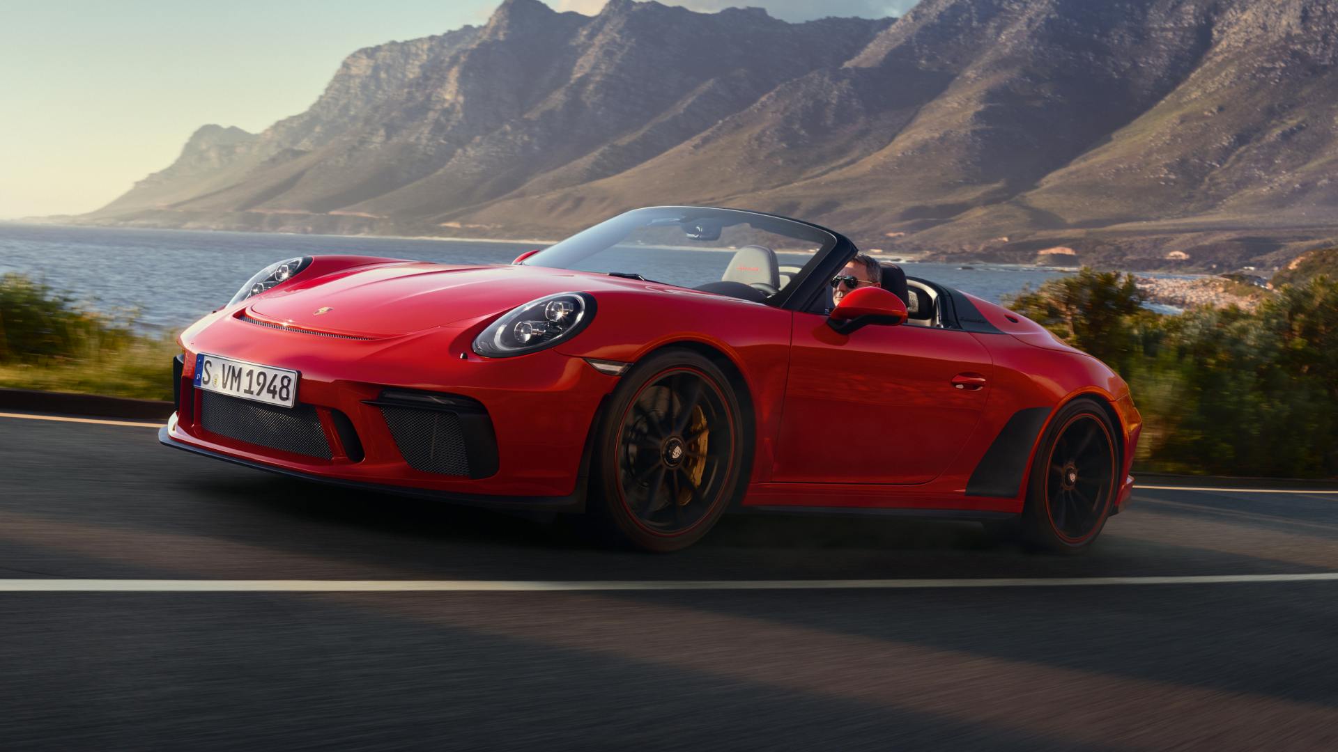 A red Porsche 911 Speedster driving along a coastal road with mountains and the ocean in the background under warm, natural light.