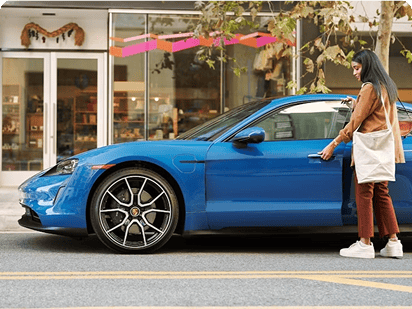 Eine Frau mit dunklen Haaren, brauner Jacke und weißer Tasche öffnet die Tür eines blauen Porsche, der am Straßenrand vor einem Geschäft mit großen Glasfronten geparkt ist.