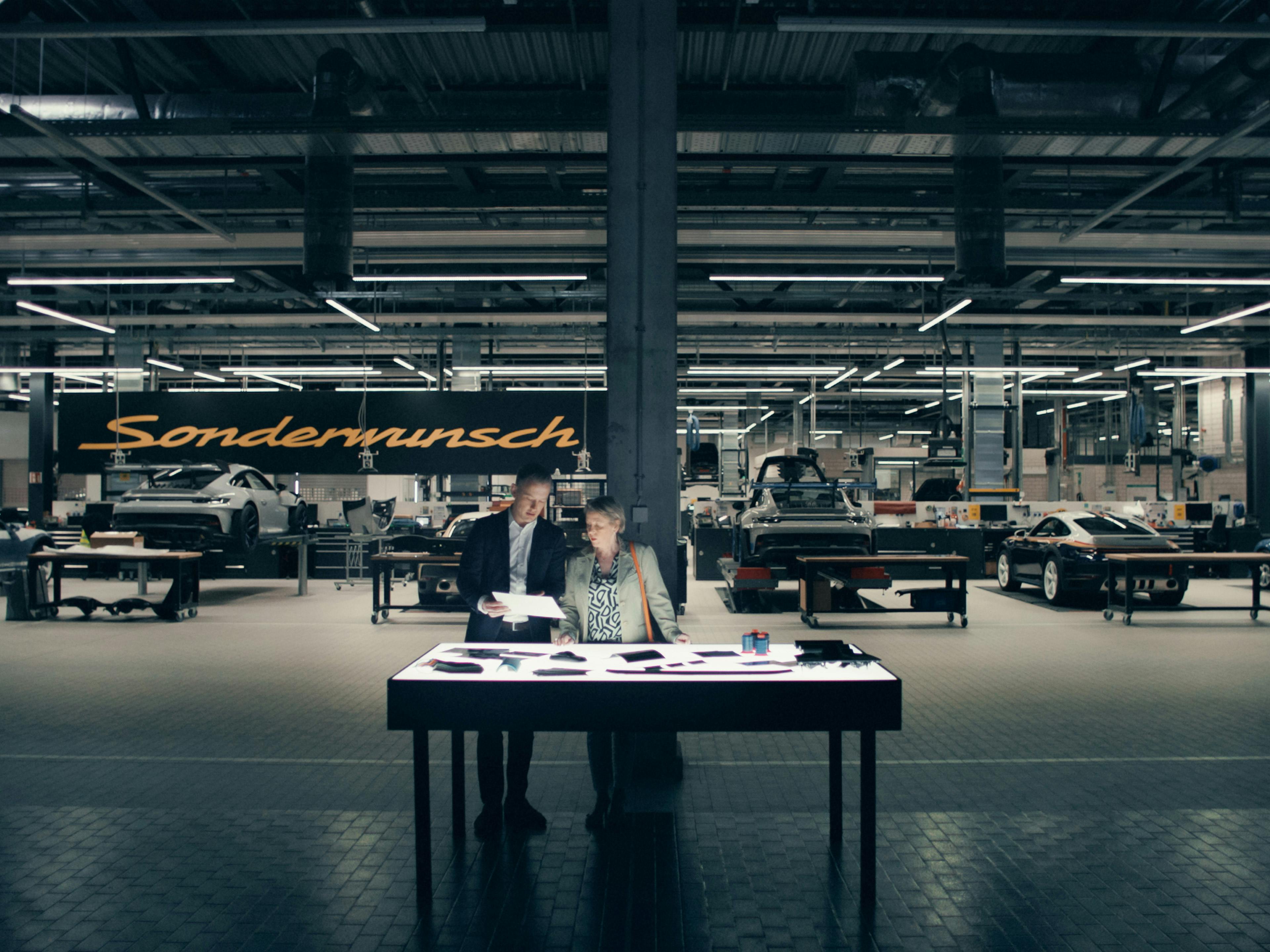 Twee mensen staan bij een tafel in Sonderwunsch Manufaktur met verschillende auto's en het bordje ‘Sonderwunsch’ zichtbaar op de achtergrond.