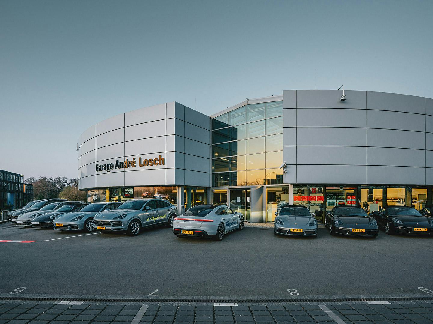Extérieur du Porsche Zenter Lëtzebuerg au crépuscule, avec de grandes lettres indiquant "Garage André Losch" au-dessus de l'entrée et plusieurs voitures Porsche stationnées devant le bâtiment.