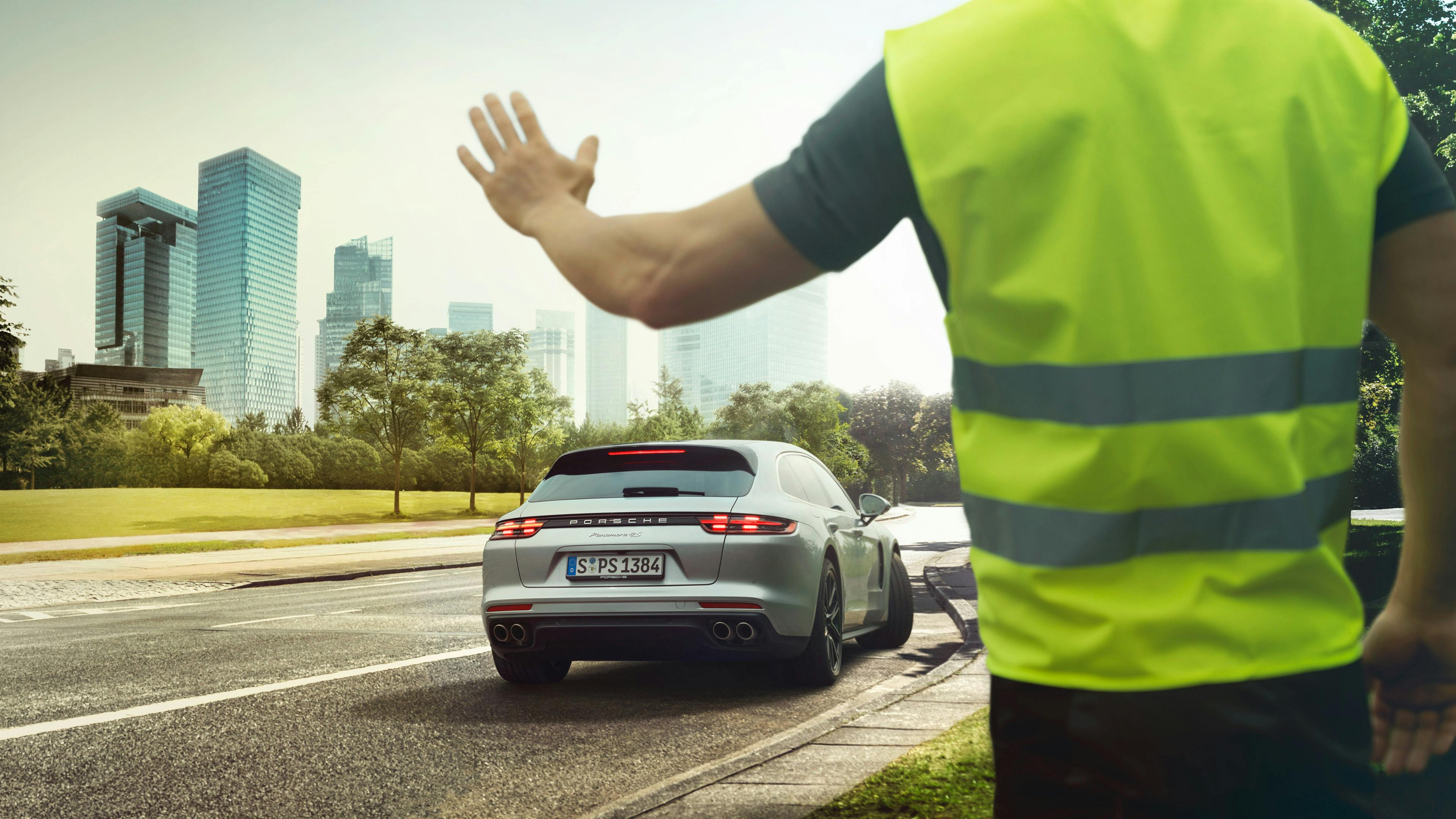 Eine Person in einer Warnweste winkt einem gerade abfahrenden, weißen Porsche Panamera in der Stadt zu.
