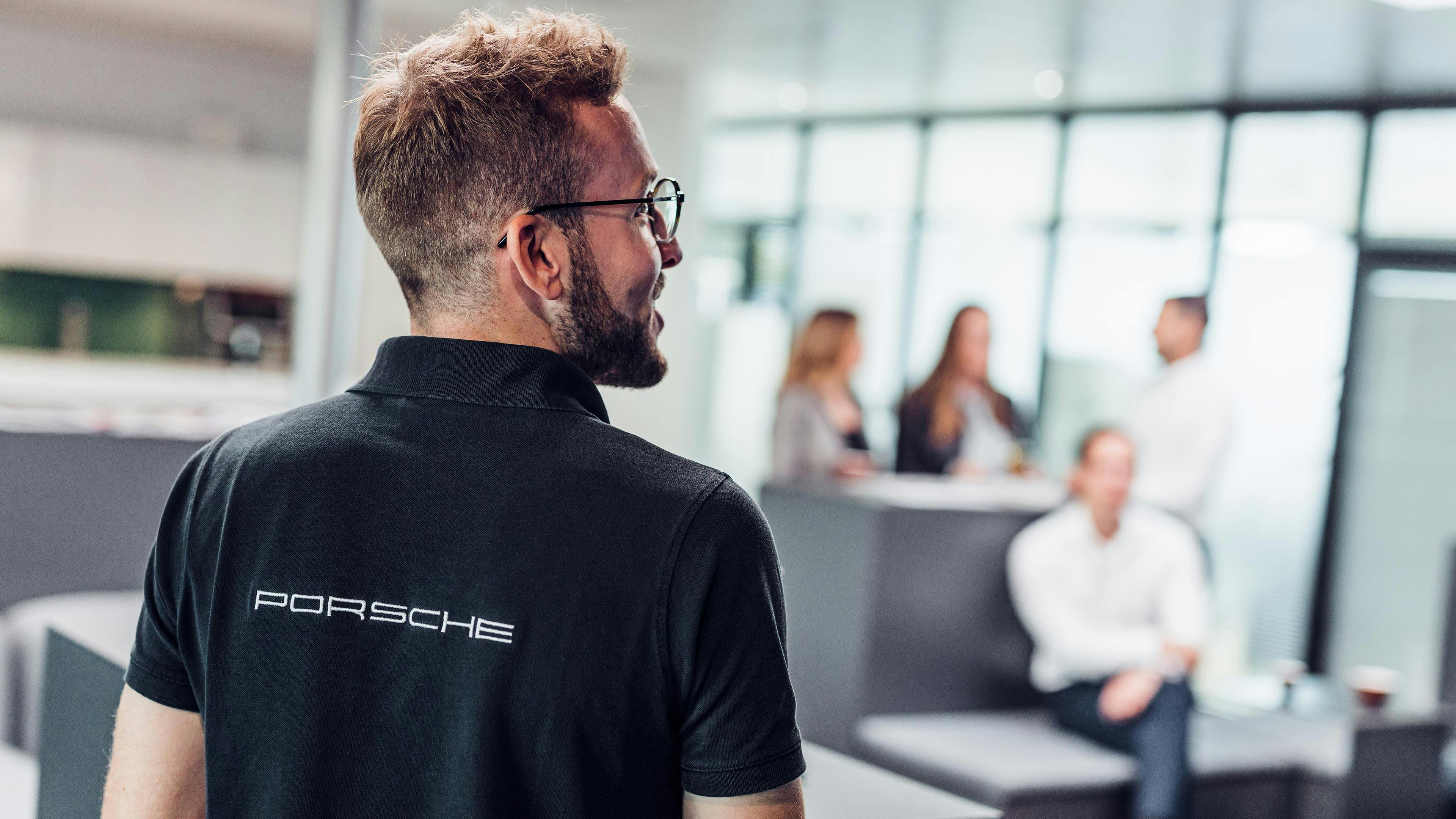 Ein Mann mit Brille in einem schwarzen Porsche T-Shirt steht mit dem Rücken zur Kamera in einem Raum mit anderen Mitarbeitern.