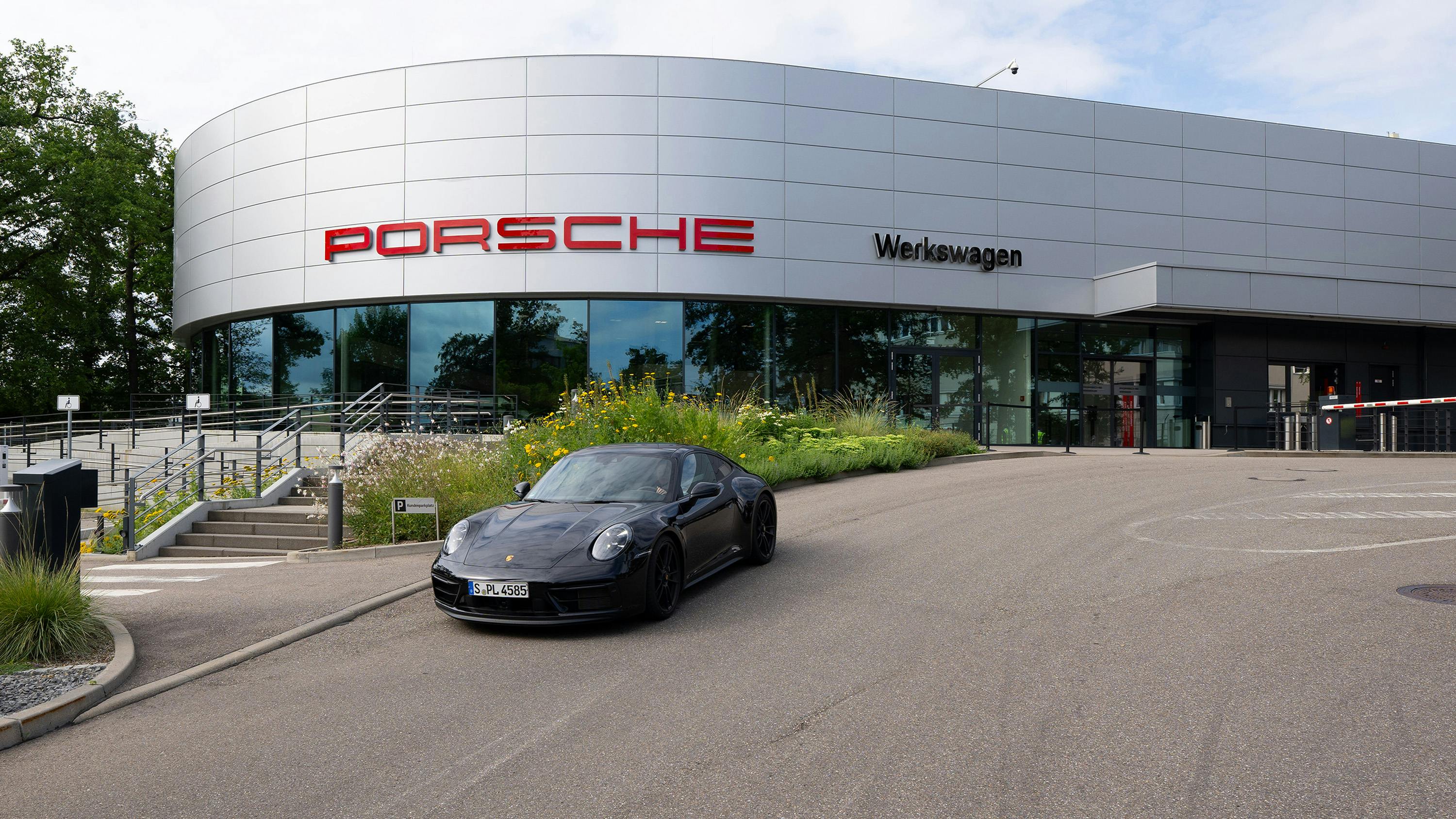 Schwarzer Porsche 911 vor modernem Werkswagen-Gebäude mit rotem Schriftzug und Glasfassade.