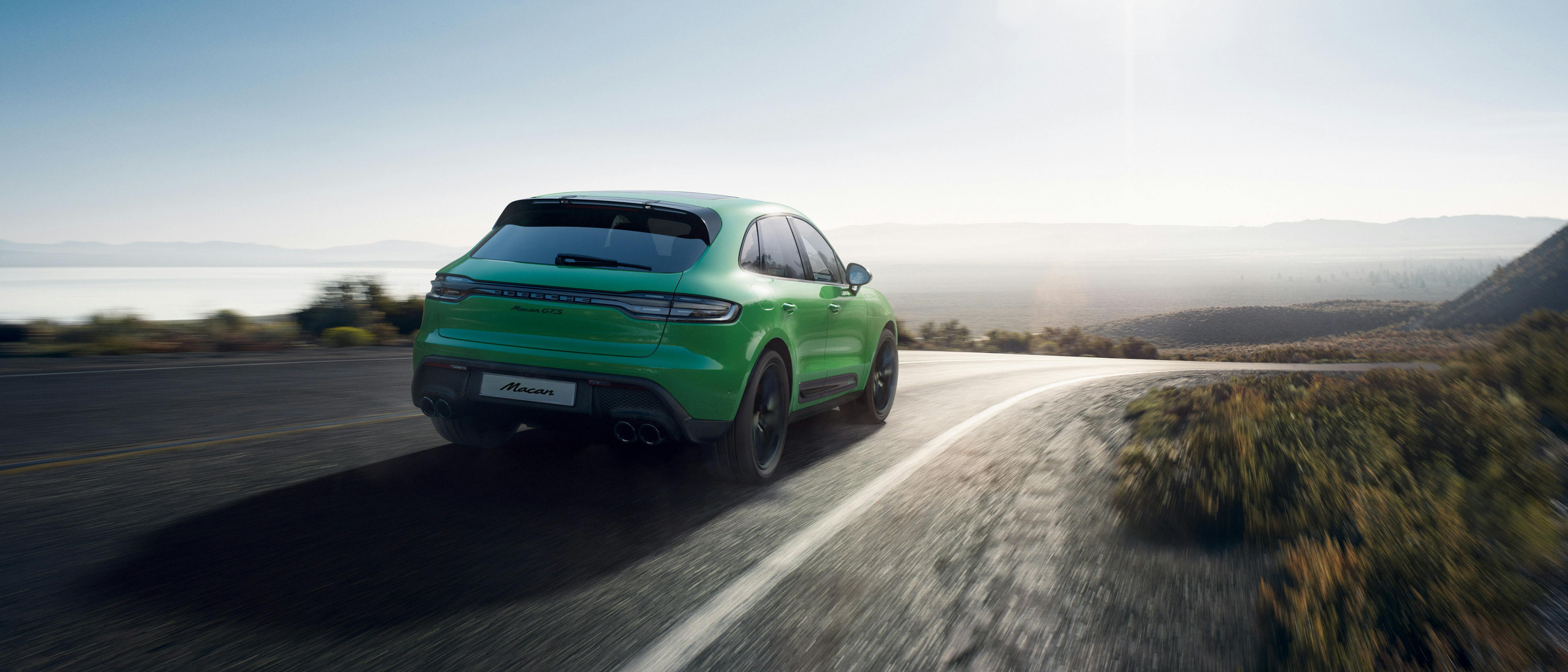 A green Porsche Macan drives on a scenic, winding road with mountains and a lake in the background under a clear sky.