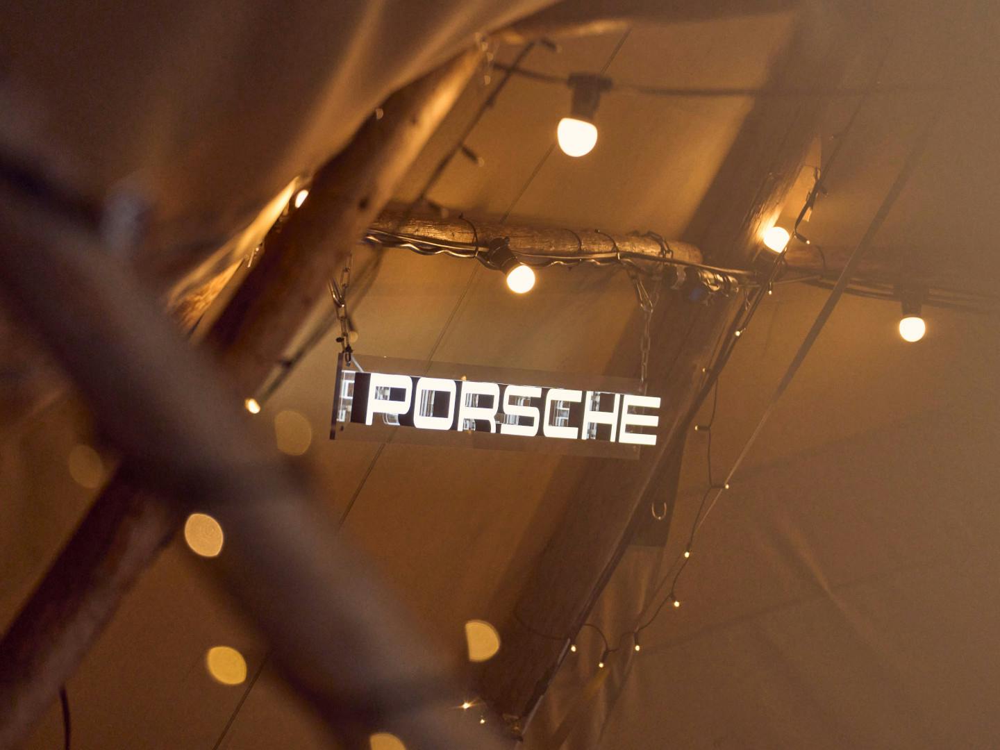 White Porsche lettering hanging from a metal structure in a tent, illuminated by an orange hue.