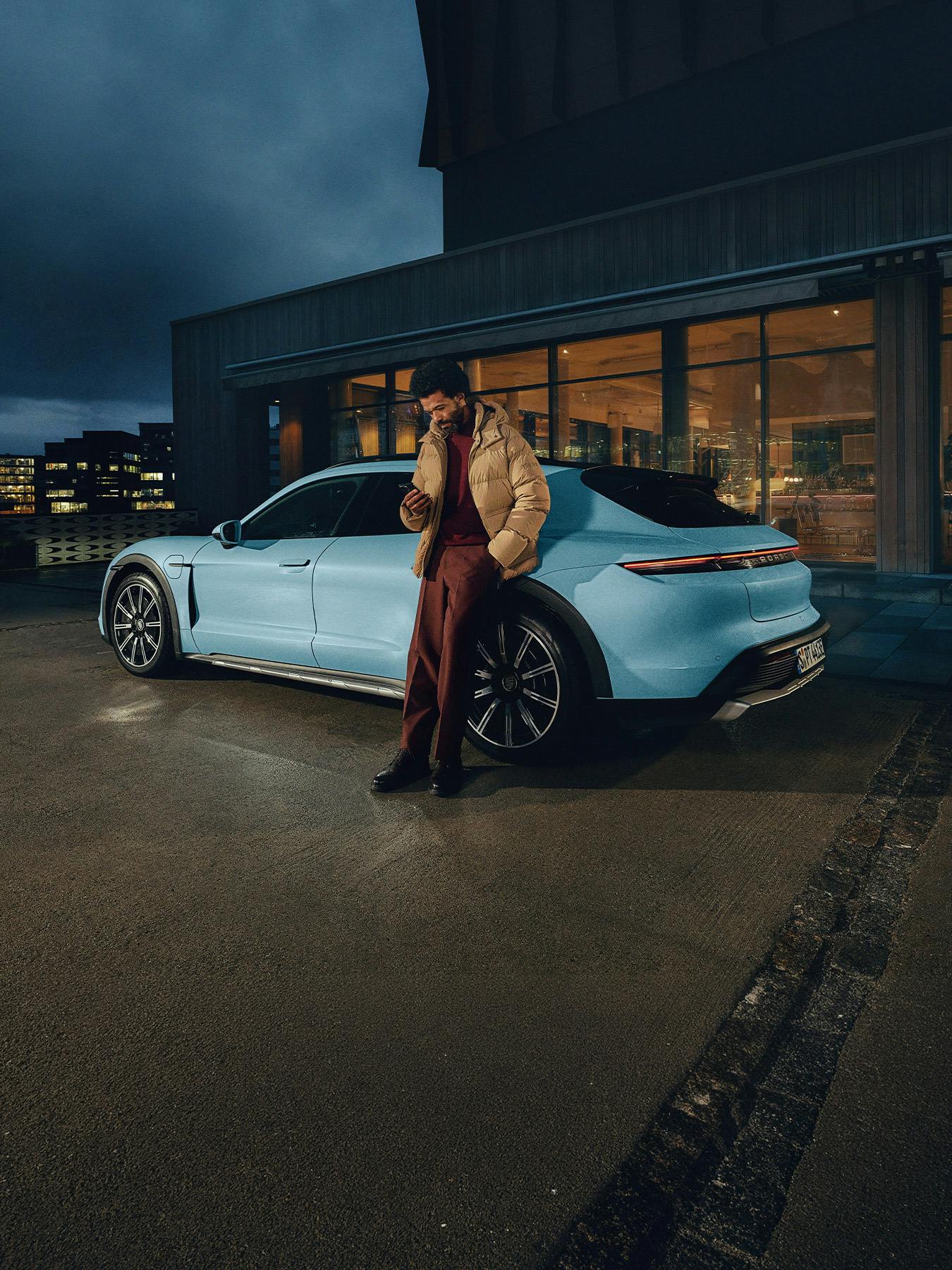 A blue Porsche Taycan standing in front of an illuminated building, with a person leaning on it.