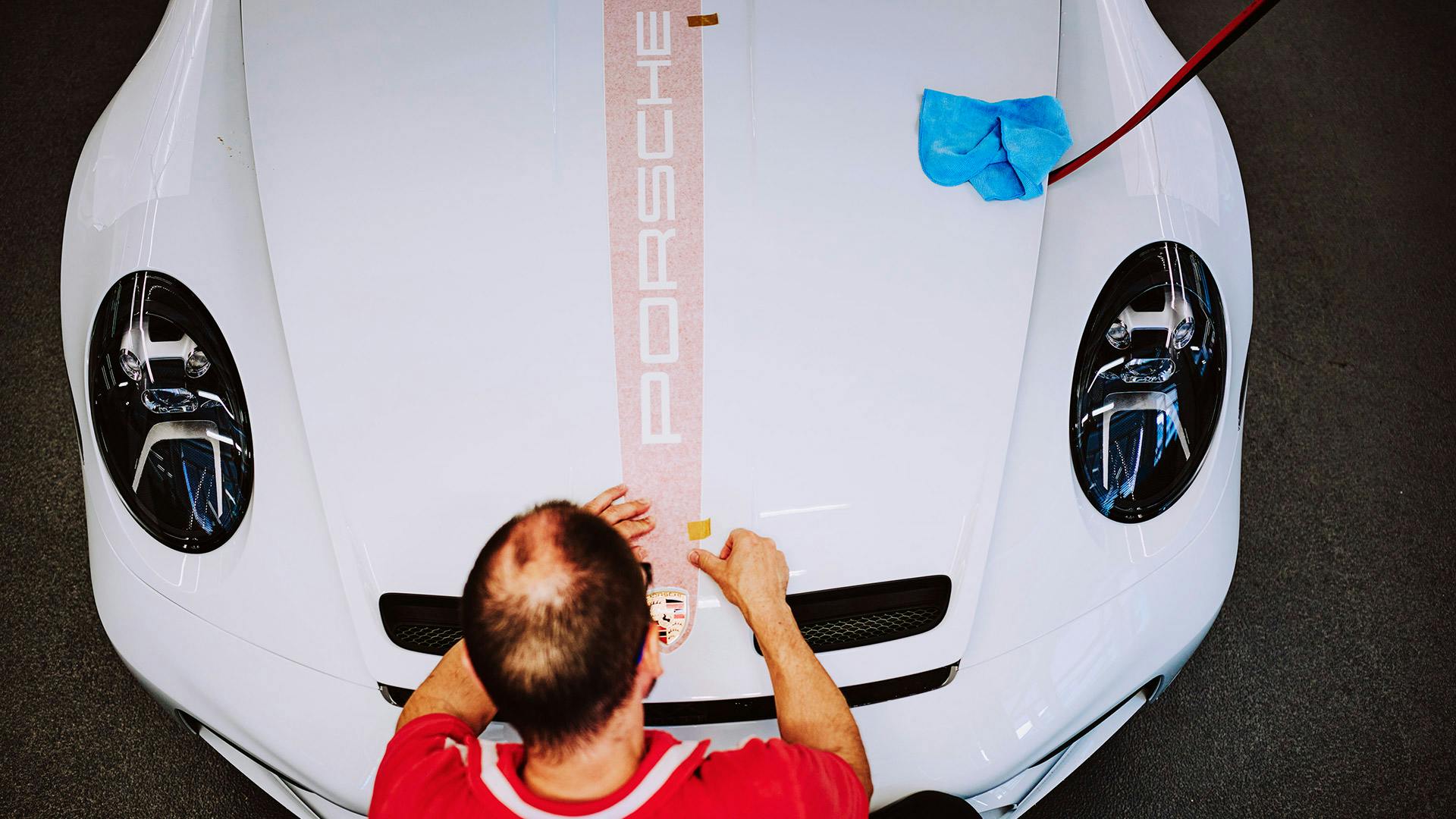 Draufsicht auf die Front eines weißen Porsche‑Sportwagens, an dem eine Person in roter Arbeitskleidung den Porsche‑Schriftzug auf der Motorhaube ausrichtet und anbringt; daneben liegen Reinigungsutensilien auf der Fahrzeugoberfläche.