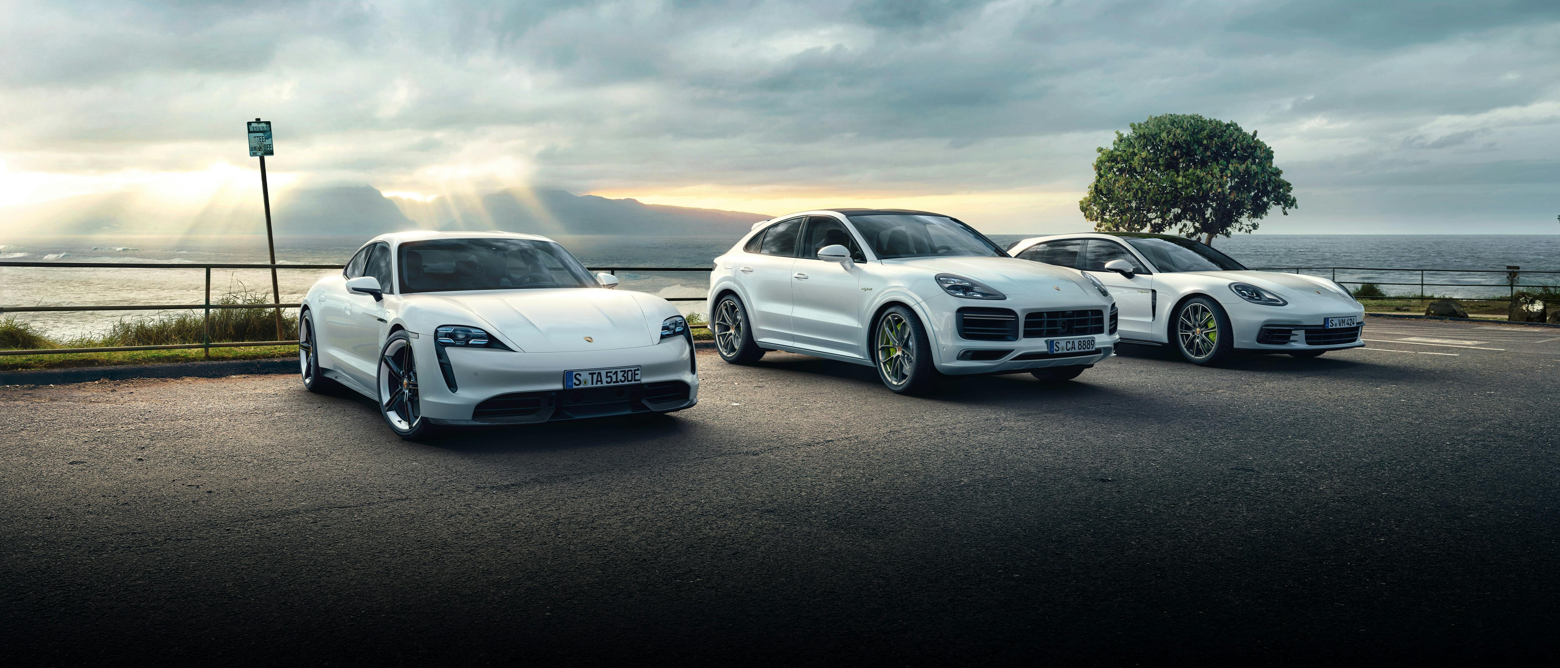 Drei weiße Porsche-Modelle (Taycan, Cayenne, Panamera) stehen nbeneinander vor einer Küstenlanschaft bei Sonnenuntergang.