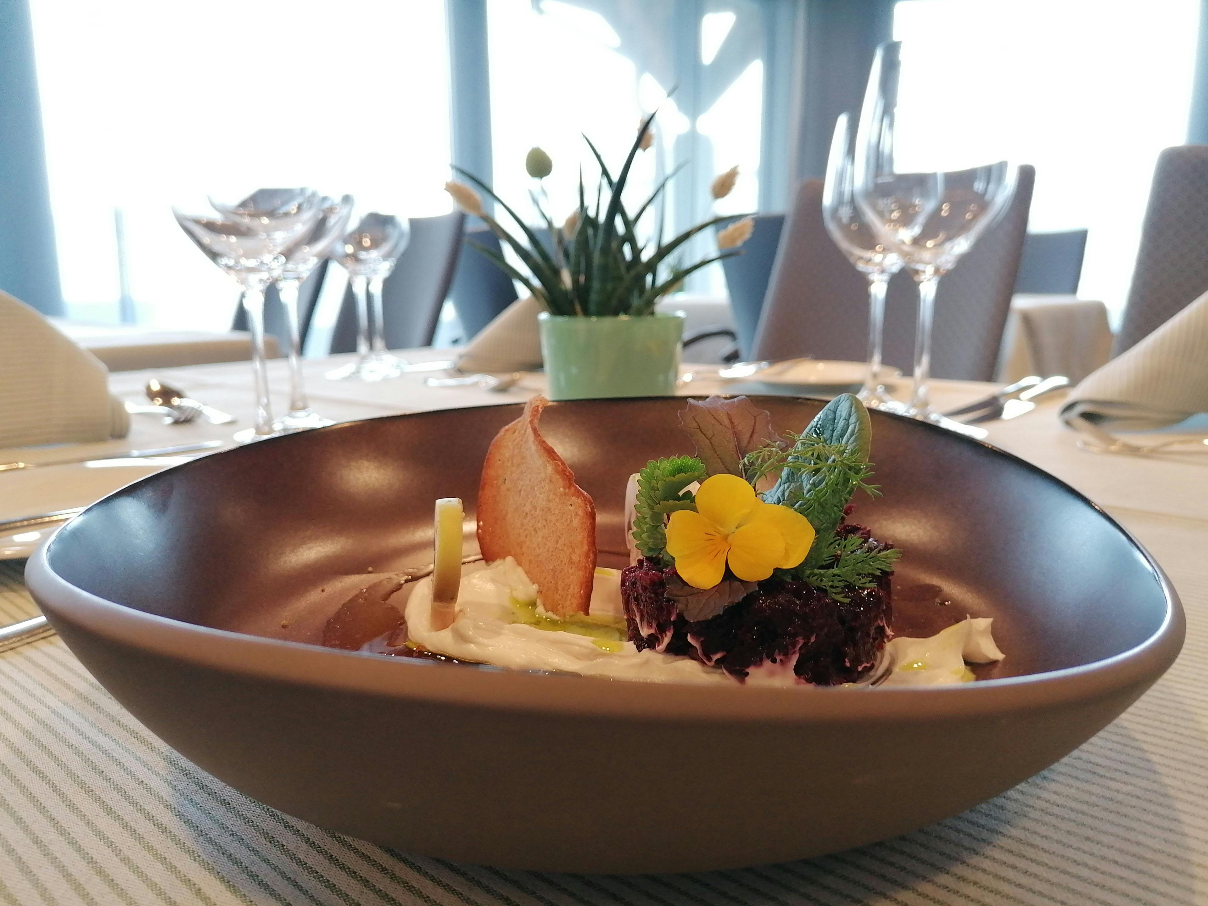 Keramikschale mit Dessert auf einem gedeckten Tisch, Weingläser und Pflanze im Hintergrund.