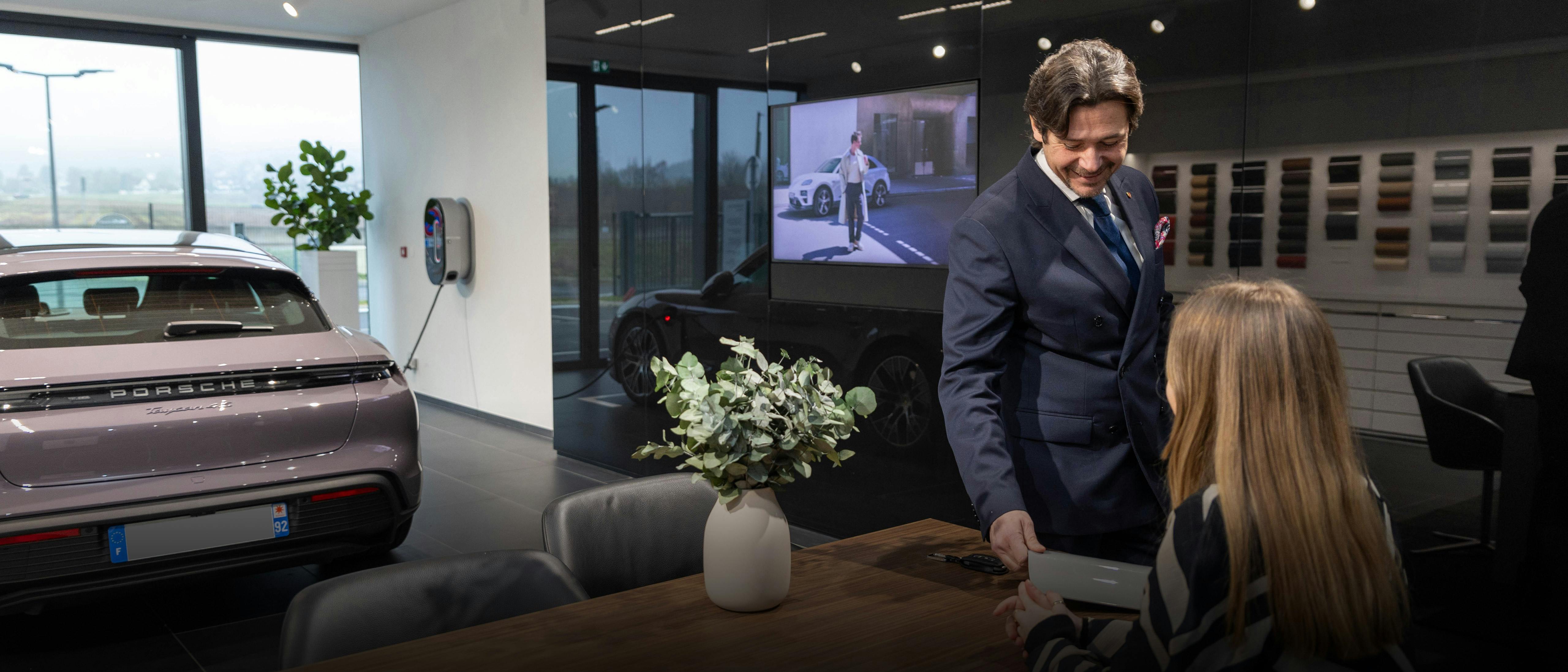 Femme assise à une table en bois dans le centre Porsche, le conseiller en entretien lui montre un échantillon de matériau gris. Mur avec la « Collection du propriétaire » en arrière-plan
