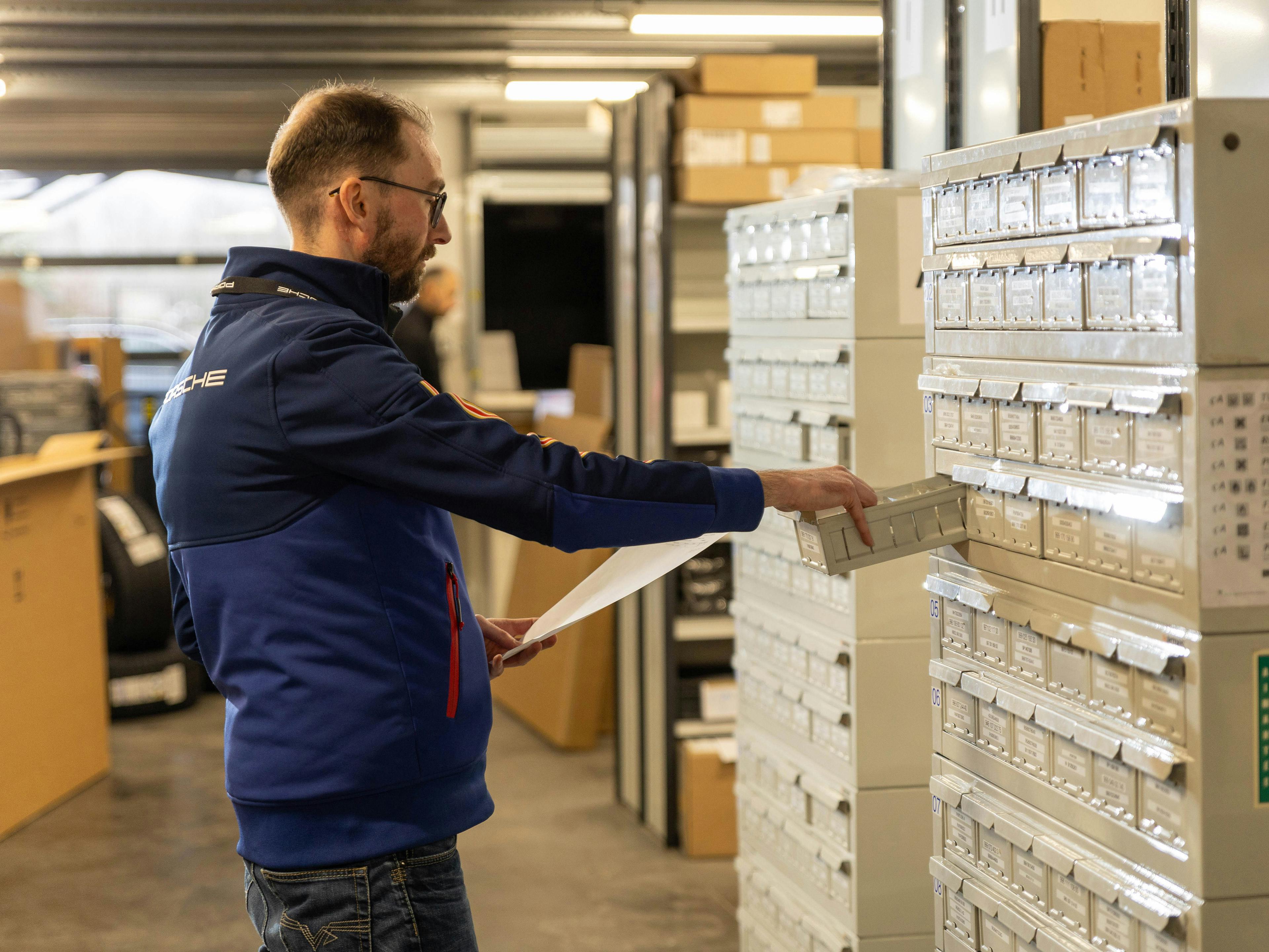 Un employé de Porsche se tient à côté d'une armoire à tiroirs et en sort un petit tiroir tout en tenant un morceau de papier dans son autre main.
