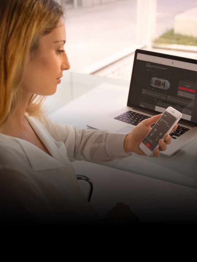 Eine Frau sitzt vor einem eingeschalteten Laptop und schaut prüfend auf das Smartphone in ihrer Hand.