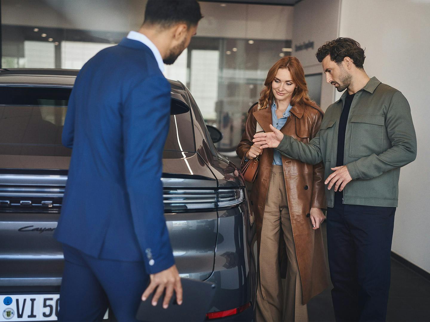 A Porsche employee in a blue suit presents a Porsche car to a couple in the showroom