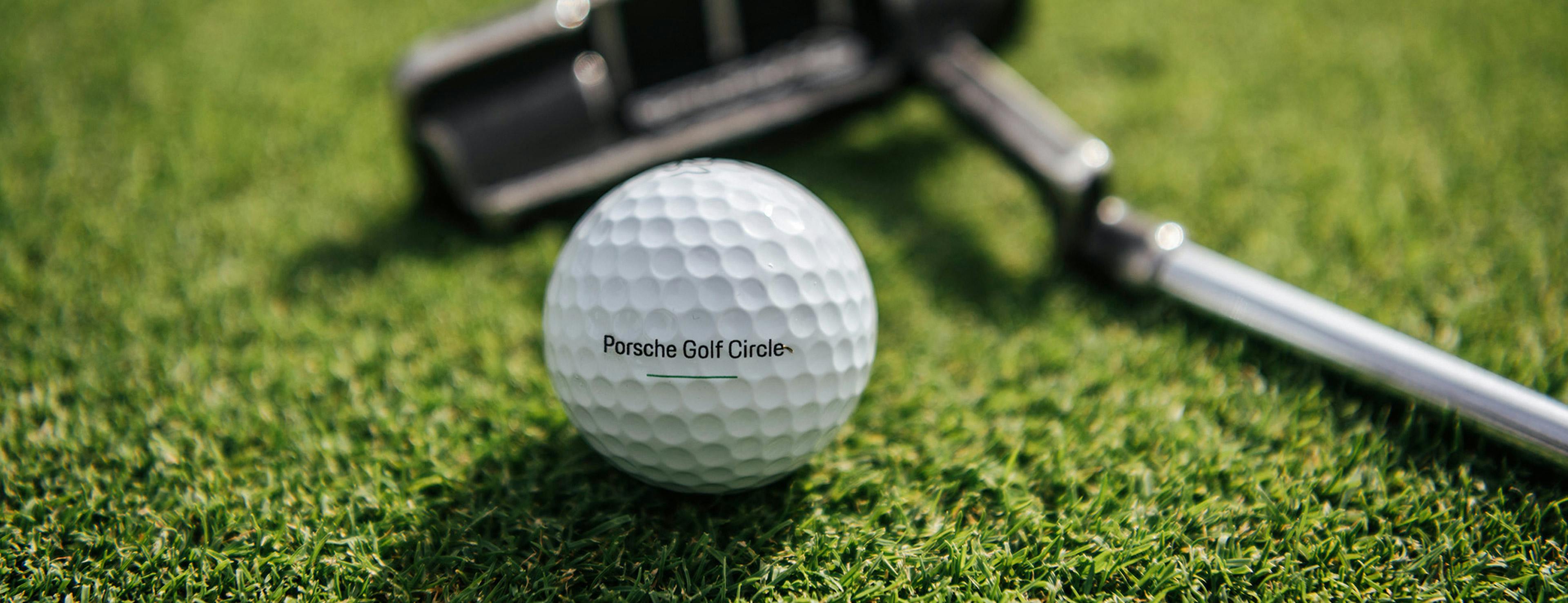 Eine Golfball mir der Aufschrift Porsche Golf Circle auf einem grünen Rasen und einem Golfschläger im Hintergrund