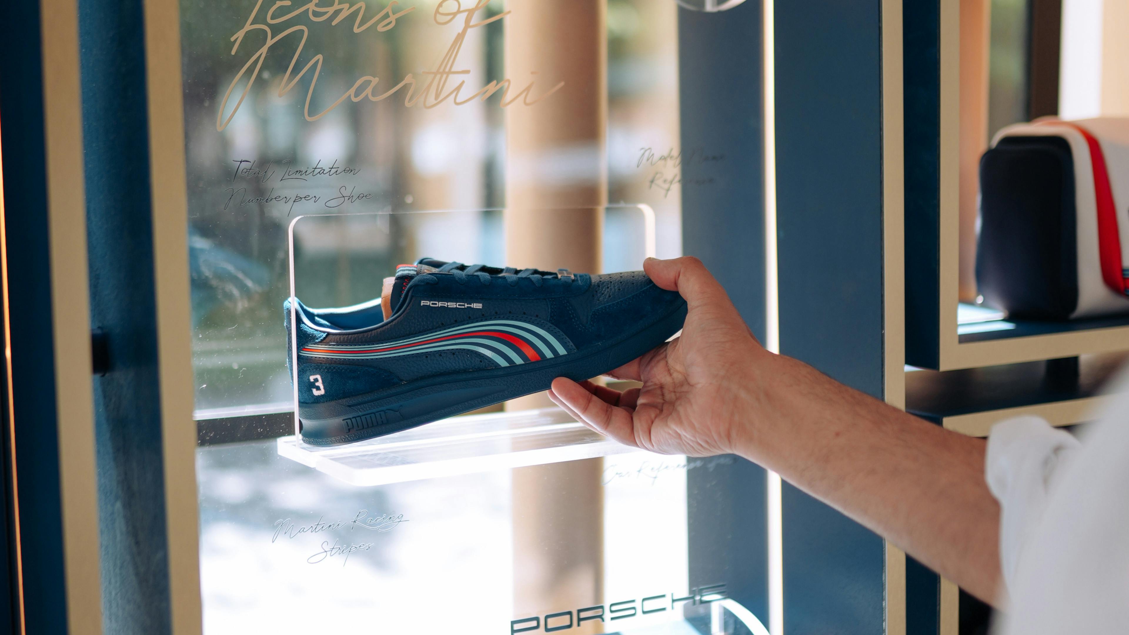 A person’s hand holds a navy-blue Puma shoe with the Martini Racing red and blue stripes. The shoe is being placed into a modern, blue display case labelled “Icons of Martini”.