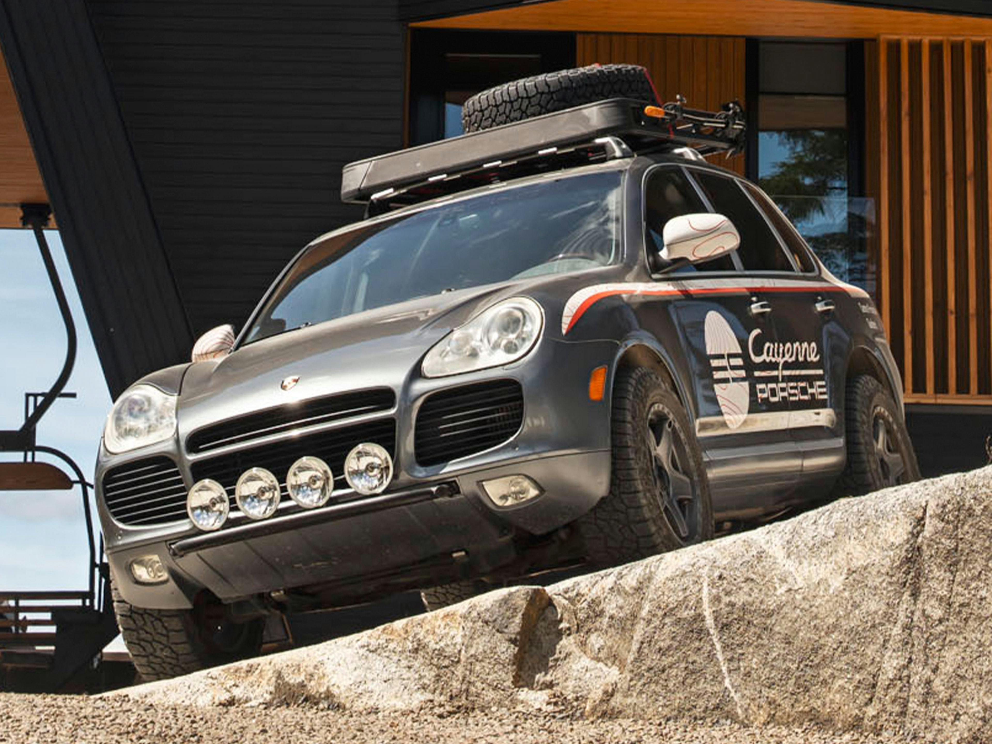 Porsche SUV with off-road equipment driving on a gravel road in a forested area.