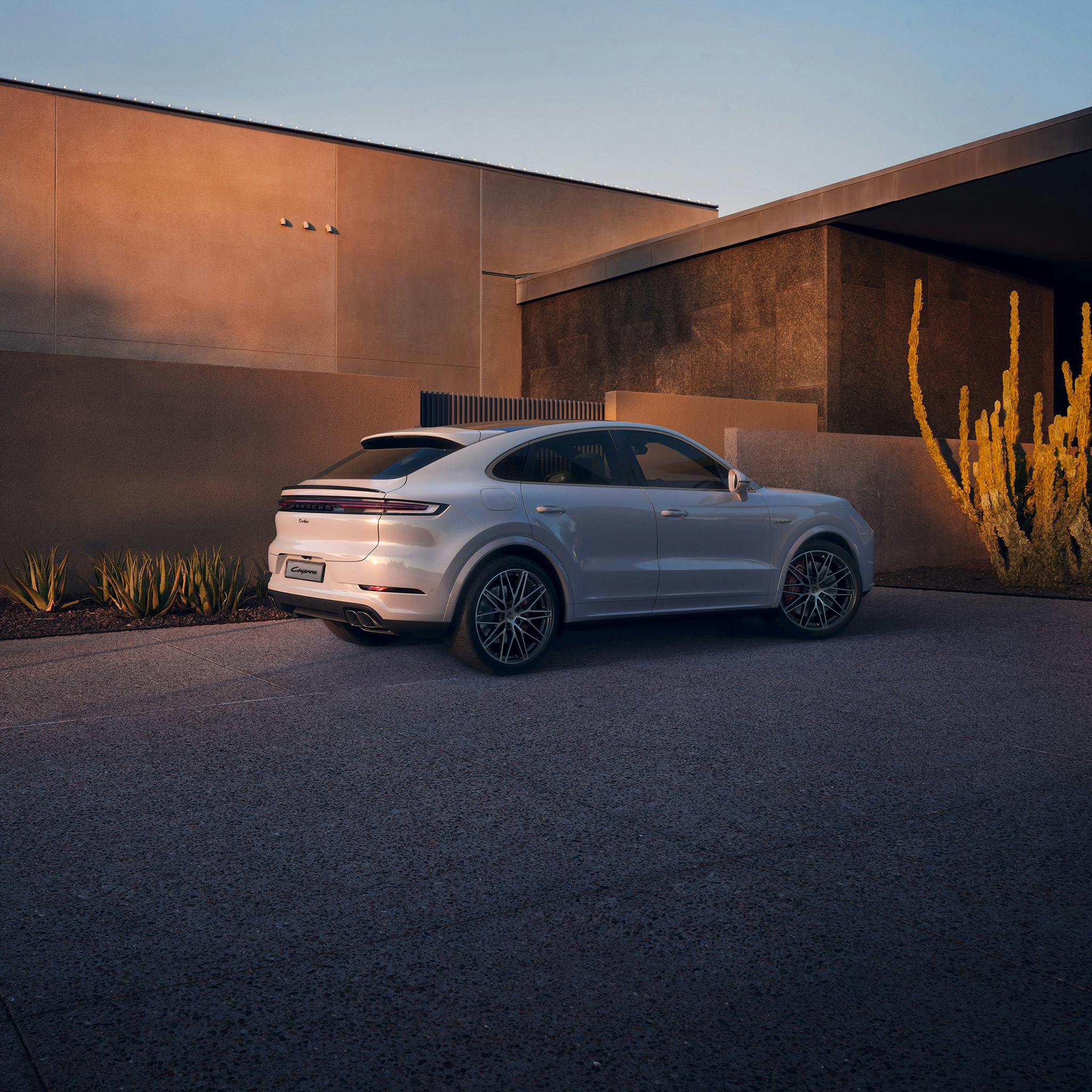 Porsche Cayenne Coupe parked in front of a modern building, surrounded by desert plants and illuminated by warm sunlight.