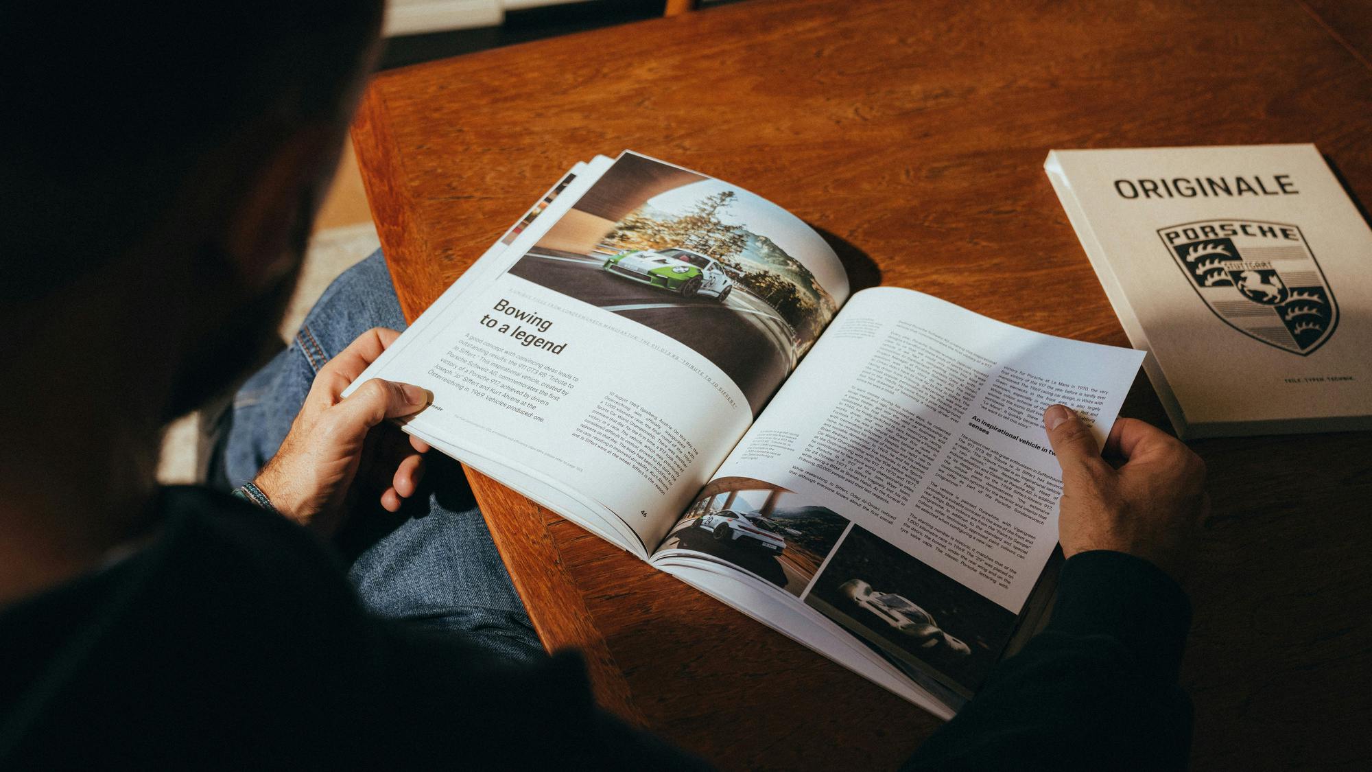 An over-the-shoulder view of a person sitting at a wooden table, reading an open car magazine. The left page features a headline that reads "Bowing to a legend" alongside a photograph of a green and white sports car driving on a road. The right page contains text and two smaller vehicle photos. To the right of the magazine, a closed tan booklet titled "ORIGINALE" sits on the table, featuring a large black Porsche crest.