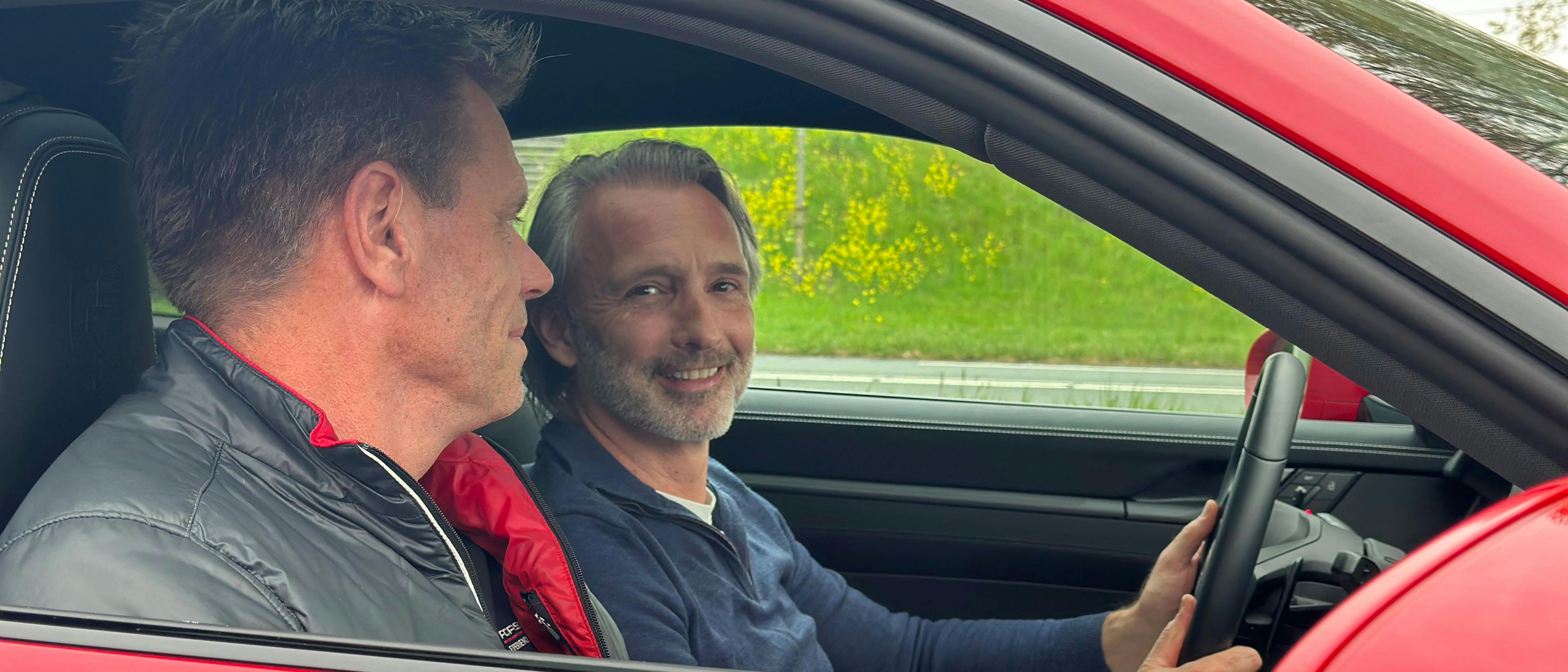 Close-up van het interieur van een rode Porsche 911 tijdens een proefrit. Een lachende man achter het stuur en een instructeur in een Porsche-bodywarmer genieten. Zij genieten van de rijervaring. De focus ligt op de luxe afwerking van het dashboard en de sportstoelen.
