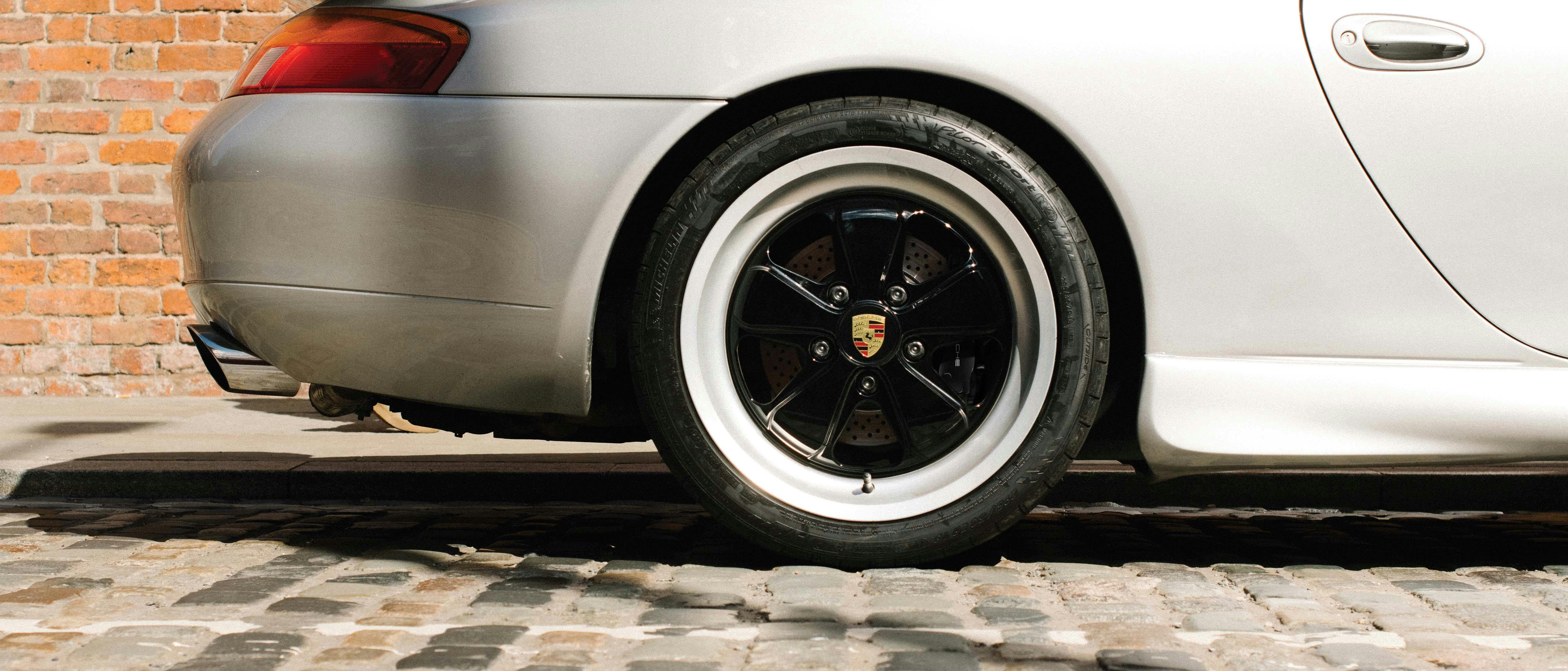 Close-up of the rear tyre of a classic silver Porsche with Fuchsfelge® (Fuchs wheel), in front of a brick wall.