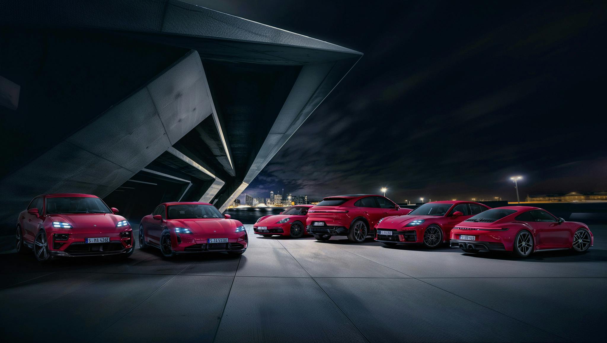Sechs rote Porsche, die nebeneinander unter einem modernen Gebäude bei Nacht geparkt sind, mit einer Stadtlandschaft im Hintergrund.