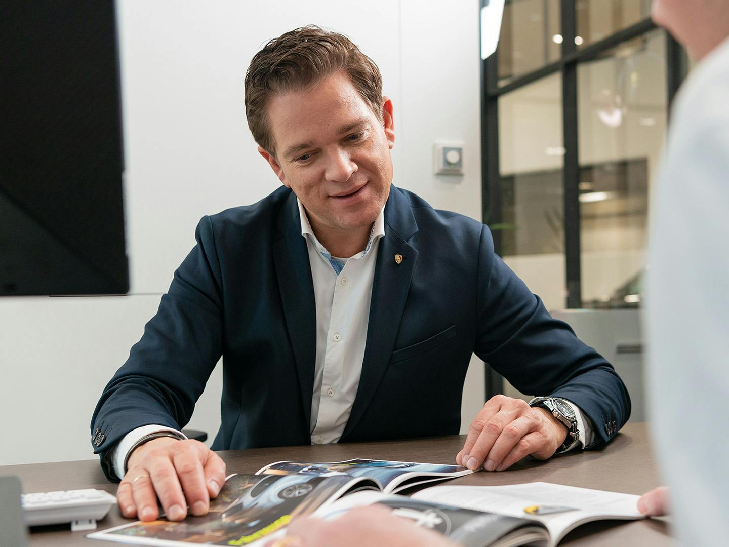 Un employé de Porsche assis derrière un bureau dans une salle de réunion montre à un collègue masculin quelque chose dans un catalogue posé sur le bureau entre eux.