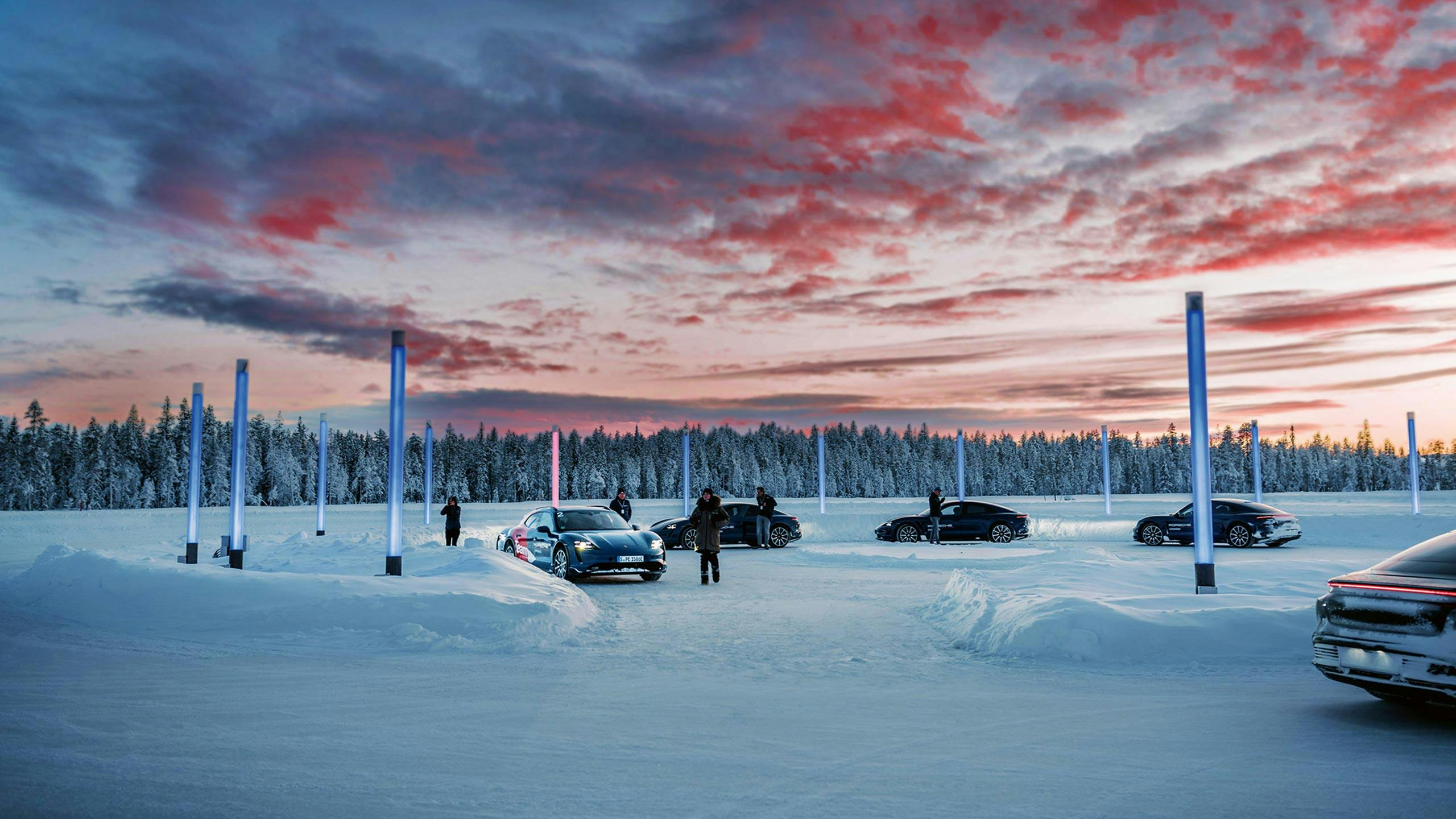 Фотография нескольких автомобилей Porsche и водителей, стоящих по кругу на заснеженной ледяной поверхности в окружении синих светящихся колонн на фоне заснеженного хвойного леса, во время мероприятия Porsche Ice Experience.