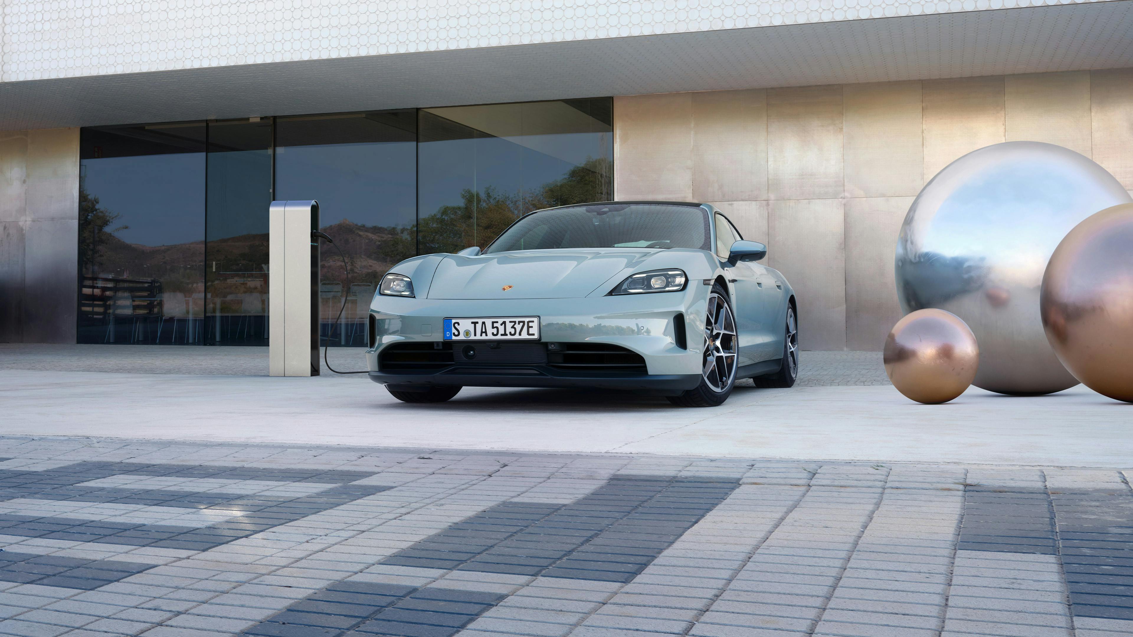 Vehículo Porsche Taycan, estacionado junto a una estación de carga frente a un edificio moderno. A la derecha, se observan esculturas esféricas con acabado metálico.