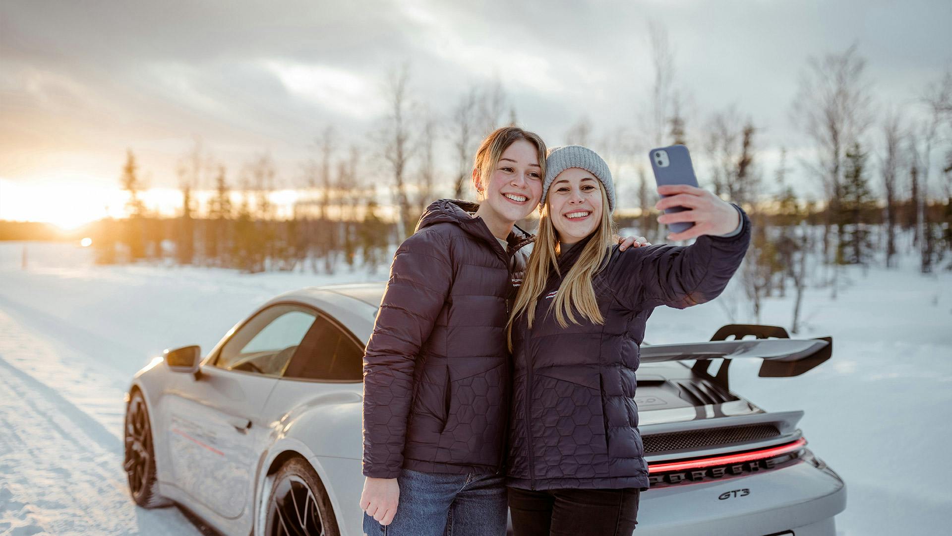 Dos participantes de Porsche Ice Experience capturan sus recuerdos con un selfie delante del vehículo y con un fondo nevado.