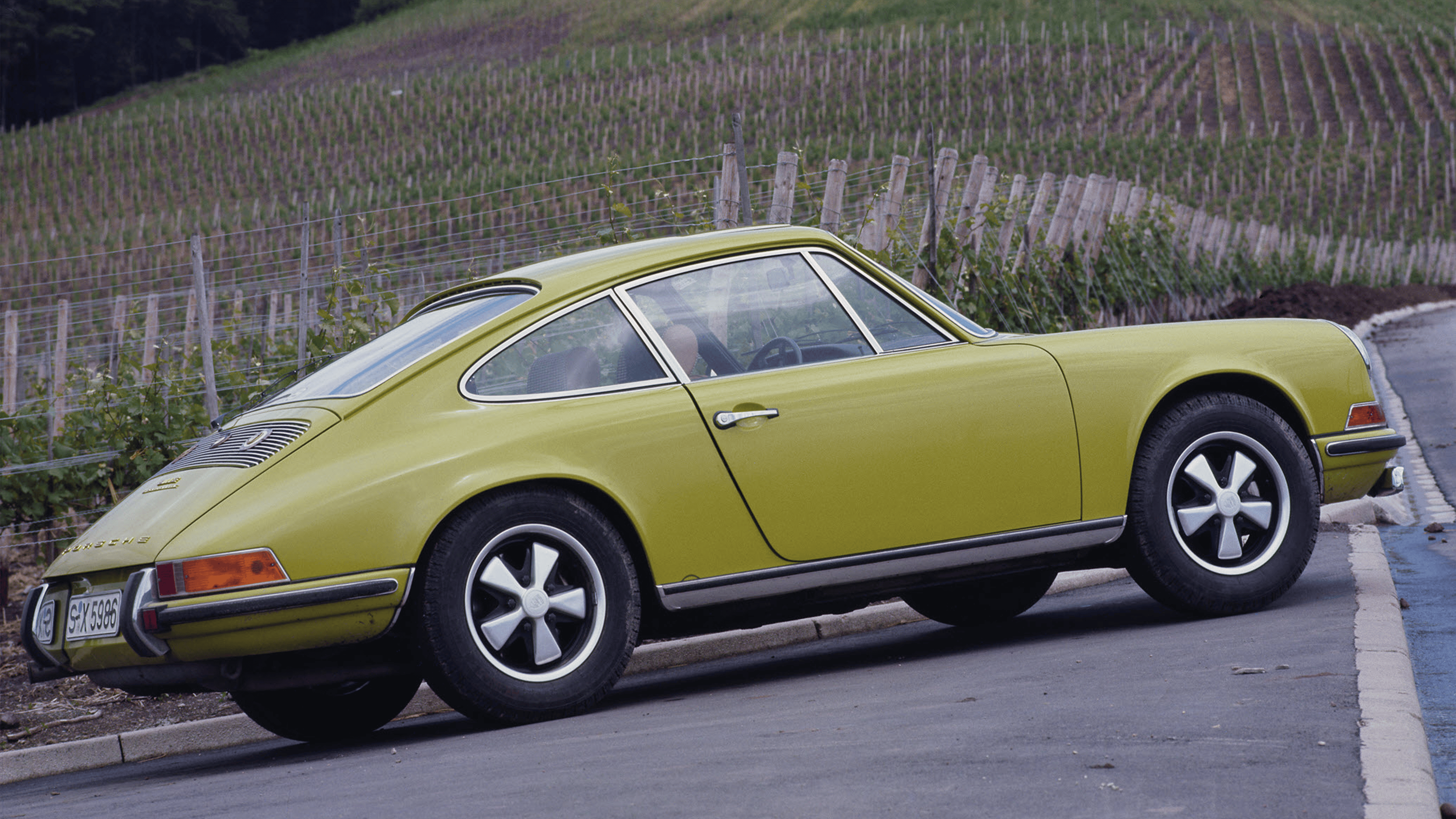 Porsche 911 E 2.2 in light green driving on the street with vineyards in the background