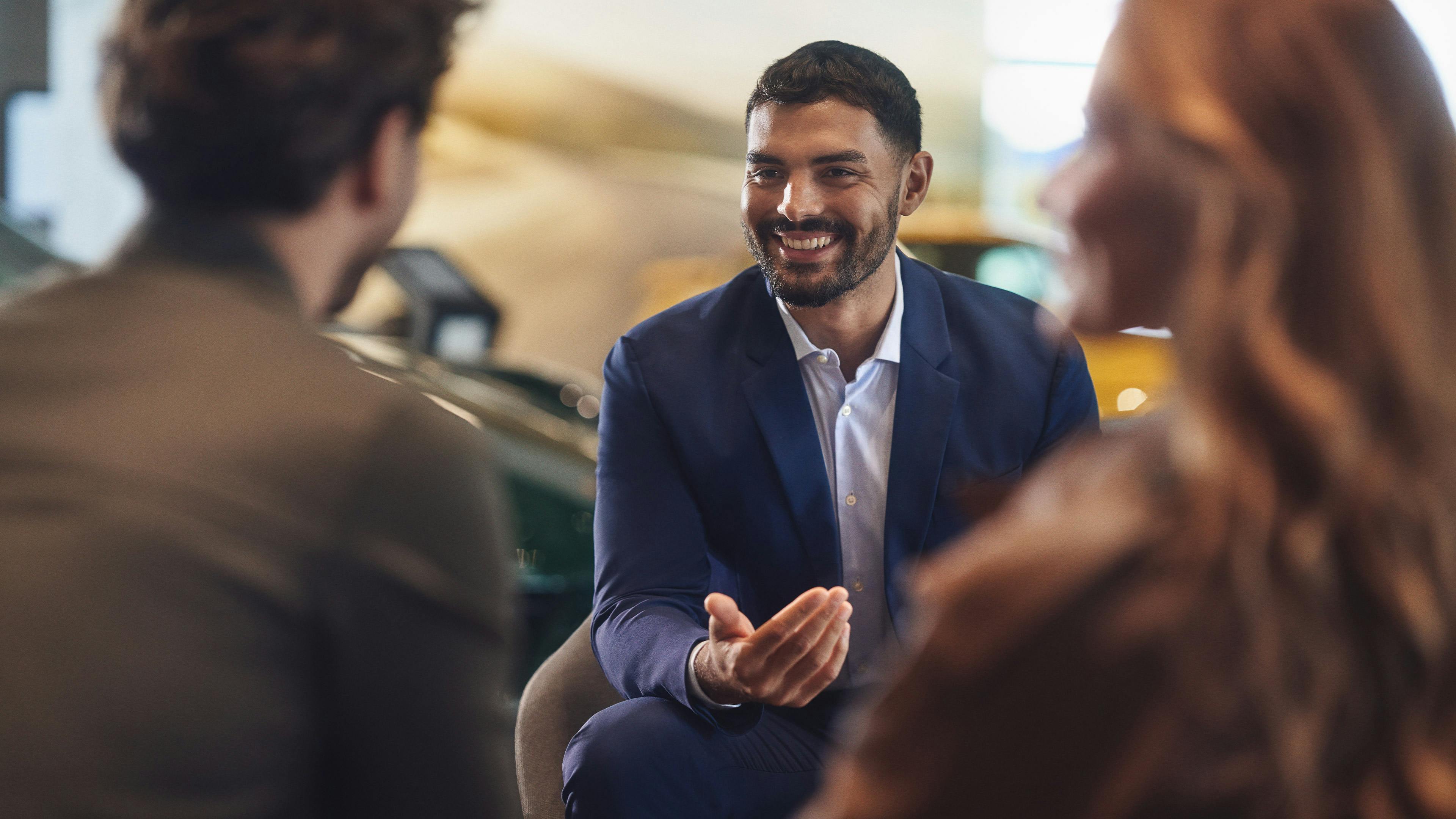 A couple and a Porsche consultant have a conversation outside a Porsche Centre. A Porsche employee and a couple are having a consultation. A Porsche can be seen in the background. In the foreground is a Mobil 1 oil bottle. A Porsche 911 can be seen in the background.