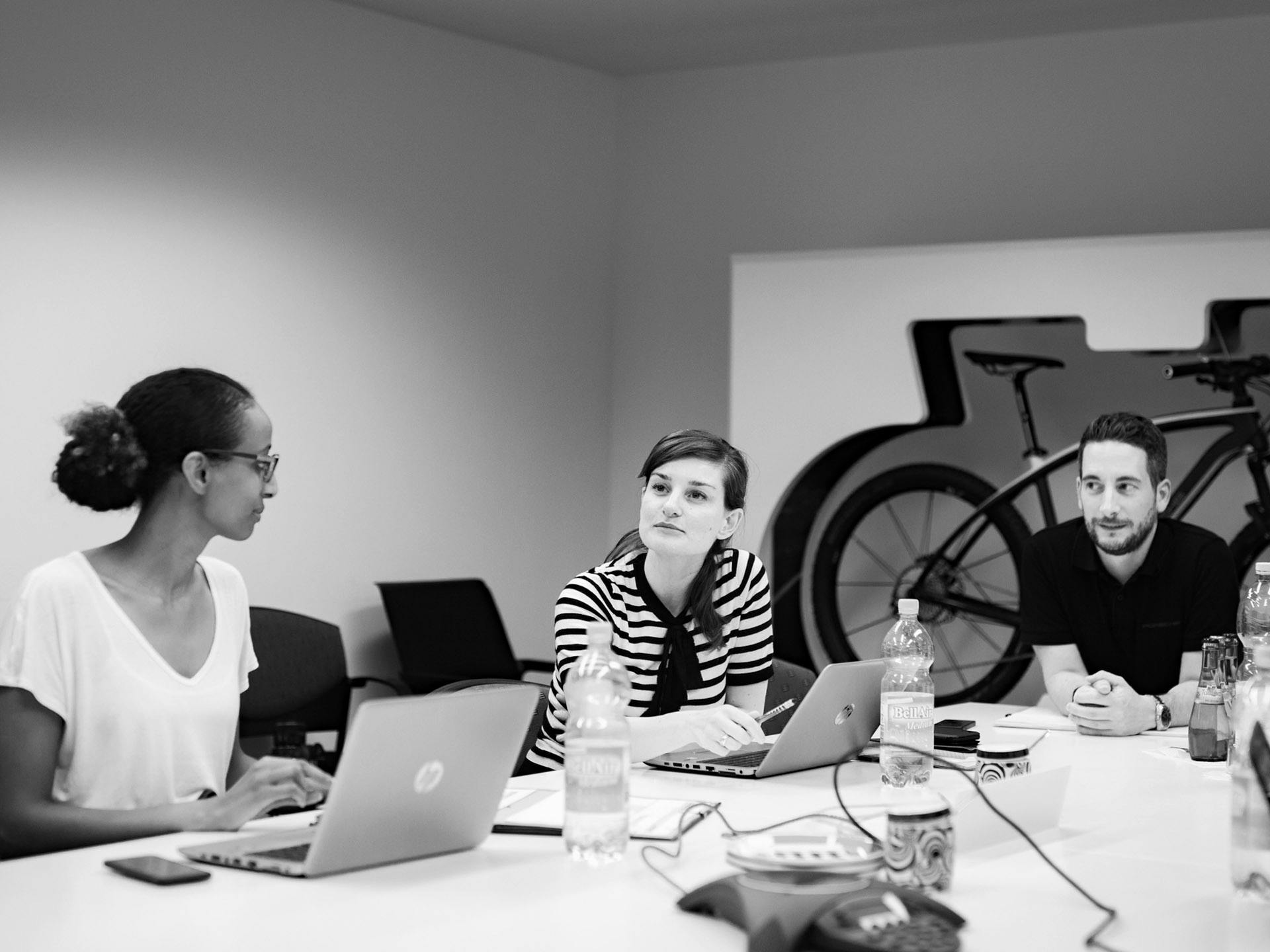 Black-and-white photo: Porsche employees sitting at a large table with laptops.