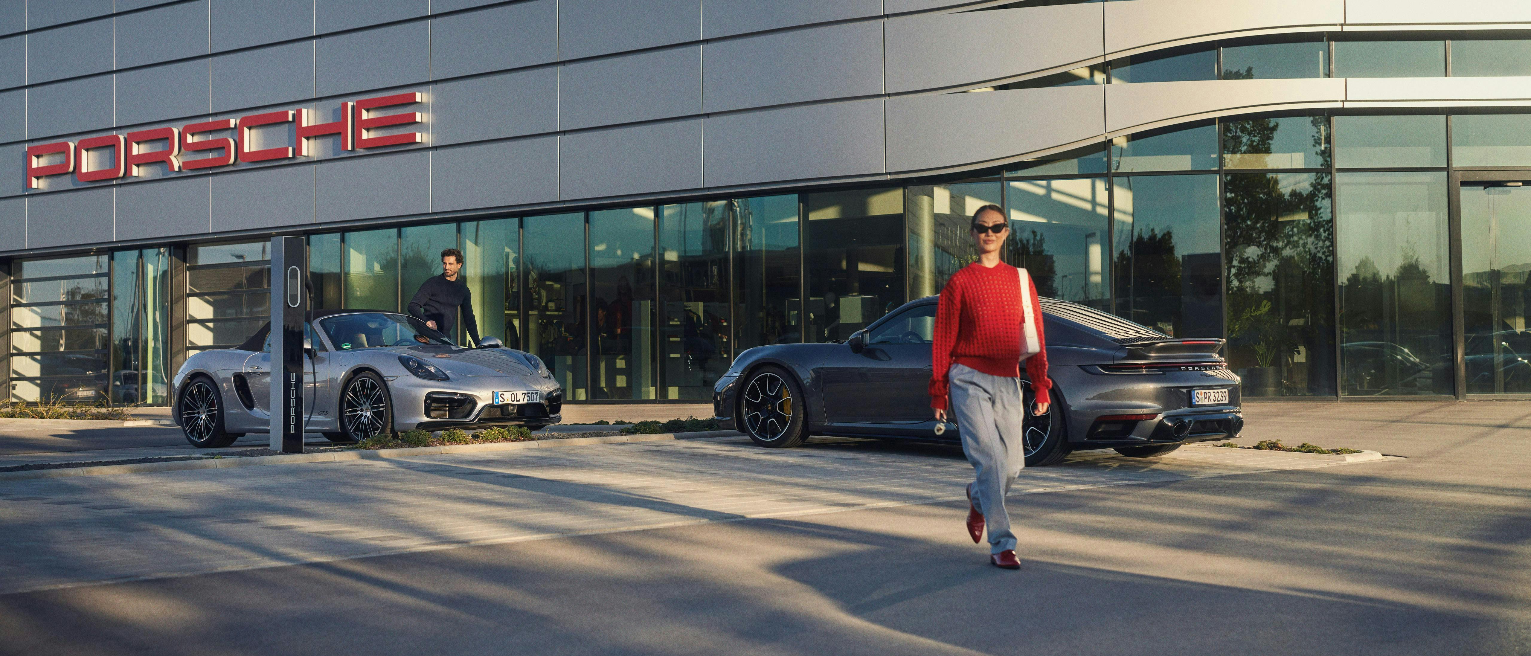 Twee mensen bij Porsche-dealer met een grijze 911 en zilveren 718 Boxster.