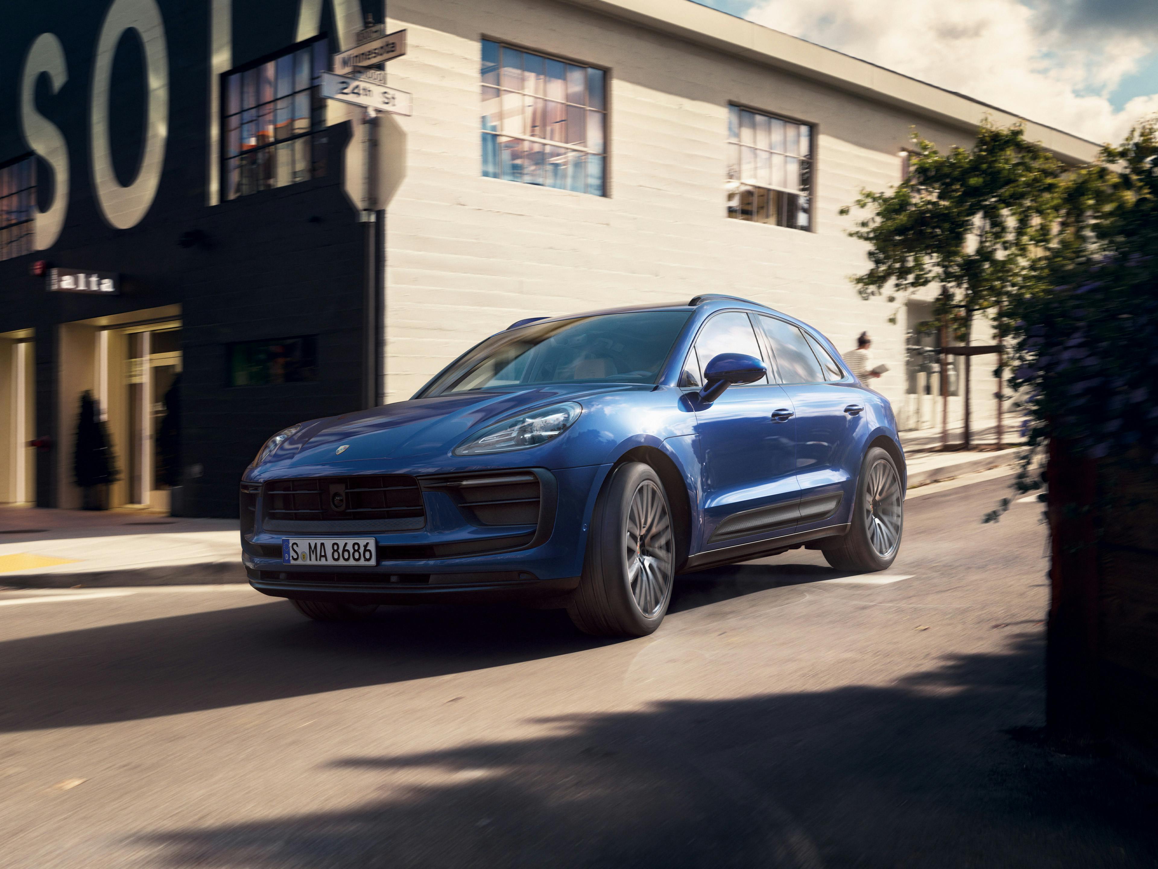The Macan driving along a road, going past a building that a white wall on one side and a brown wall on the front.