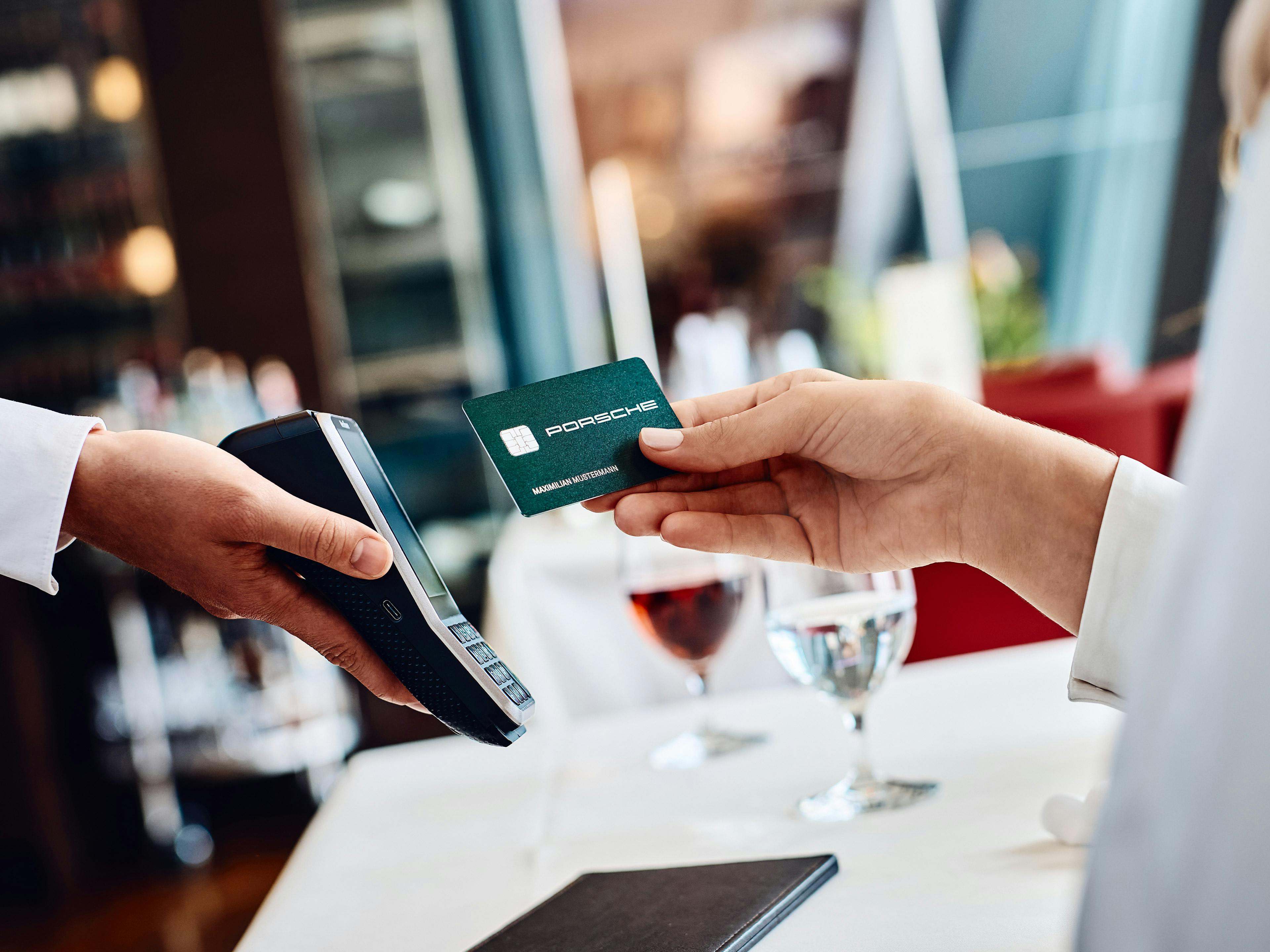 Eine Person hält eine Porsche-Kreditkarte über ein Kartenlesegerät, um eine Zahlung in einem Restaurant durchzuführen. Auf dem Tisch stehen zwei Gläser mit Getränken und eine schwarze Rechnungsmappe.
