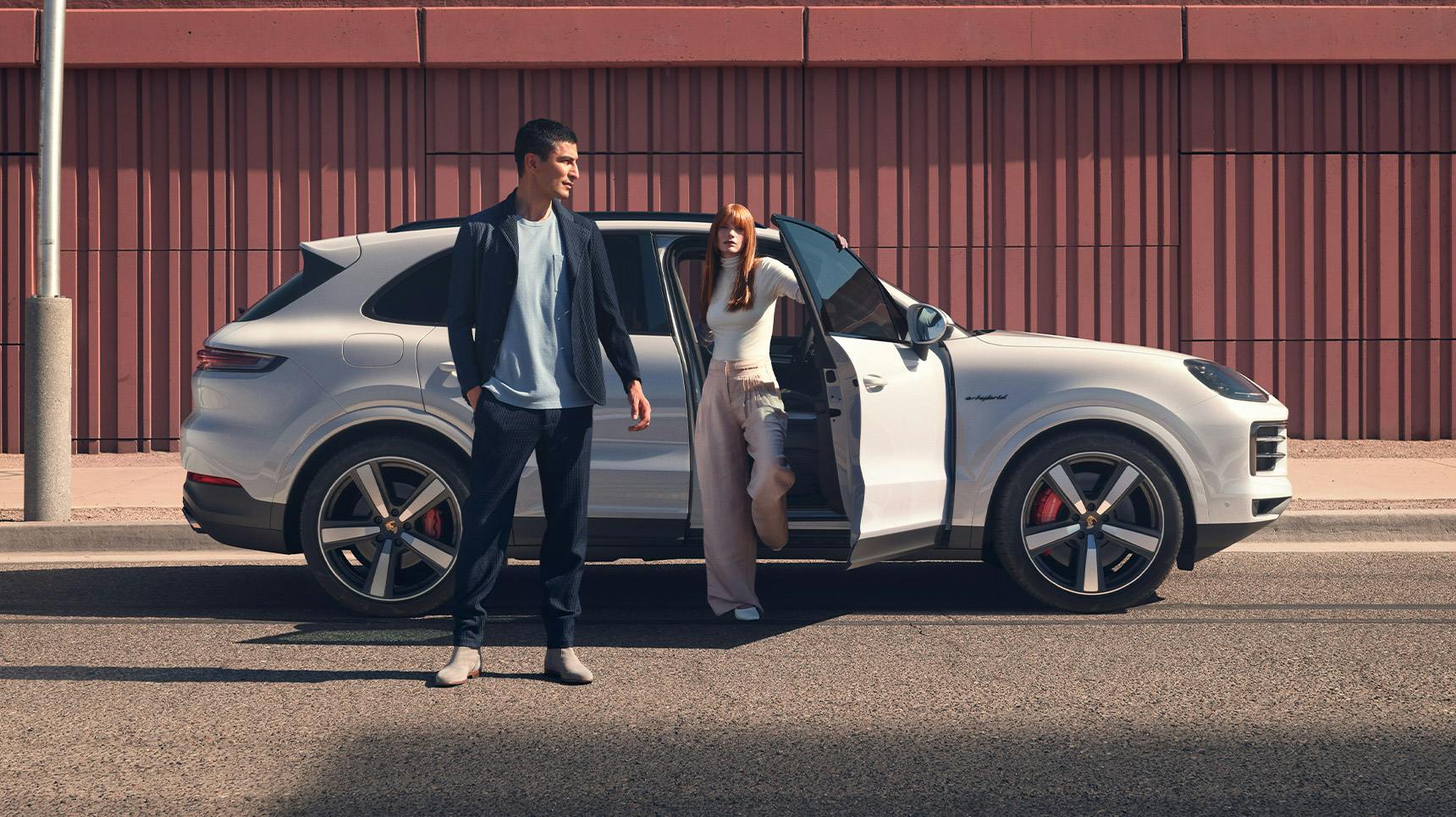 Two people stand beside a white Porsche Cayenne Turbo E-Hybrid, one exiting the vehicle near a modern building.
