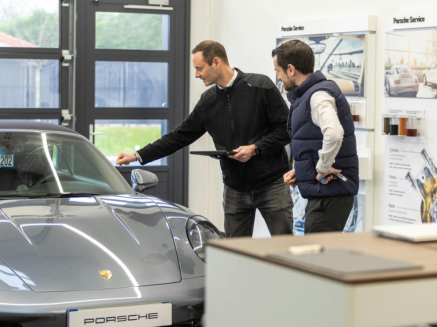 Un employé de Porsche, une tablette à la main, se tient à côté d'une Porsche grise et la désigne de l'autre main. Son collègue, debout à côté de lui, les bras sur les hanches, observe le véhicule.