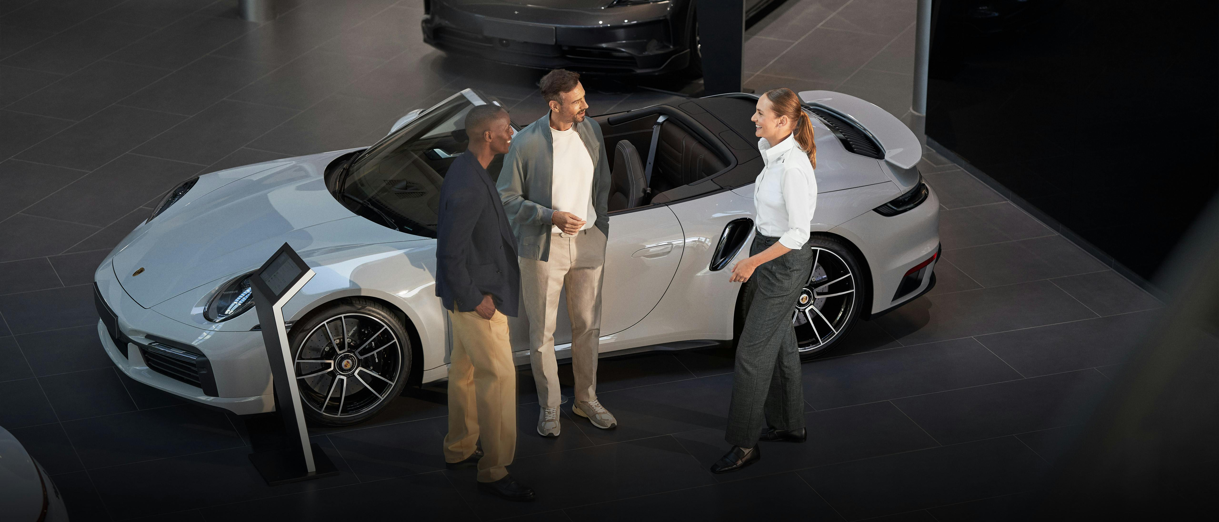 Deux hommes sont conseillés par un employé de Porsche dans la salle d'exposition devant une Porsche 911 Cabrio grise. À l'arrière-plan se trouvent deux autres Porsche.