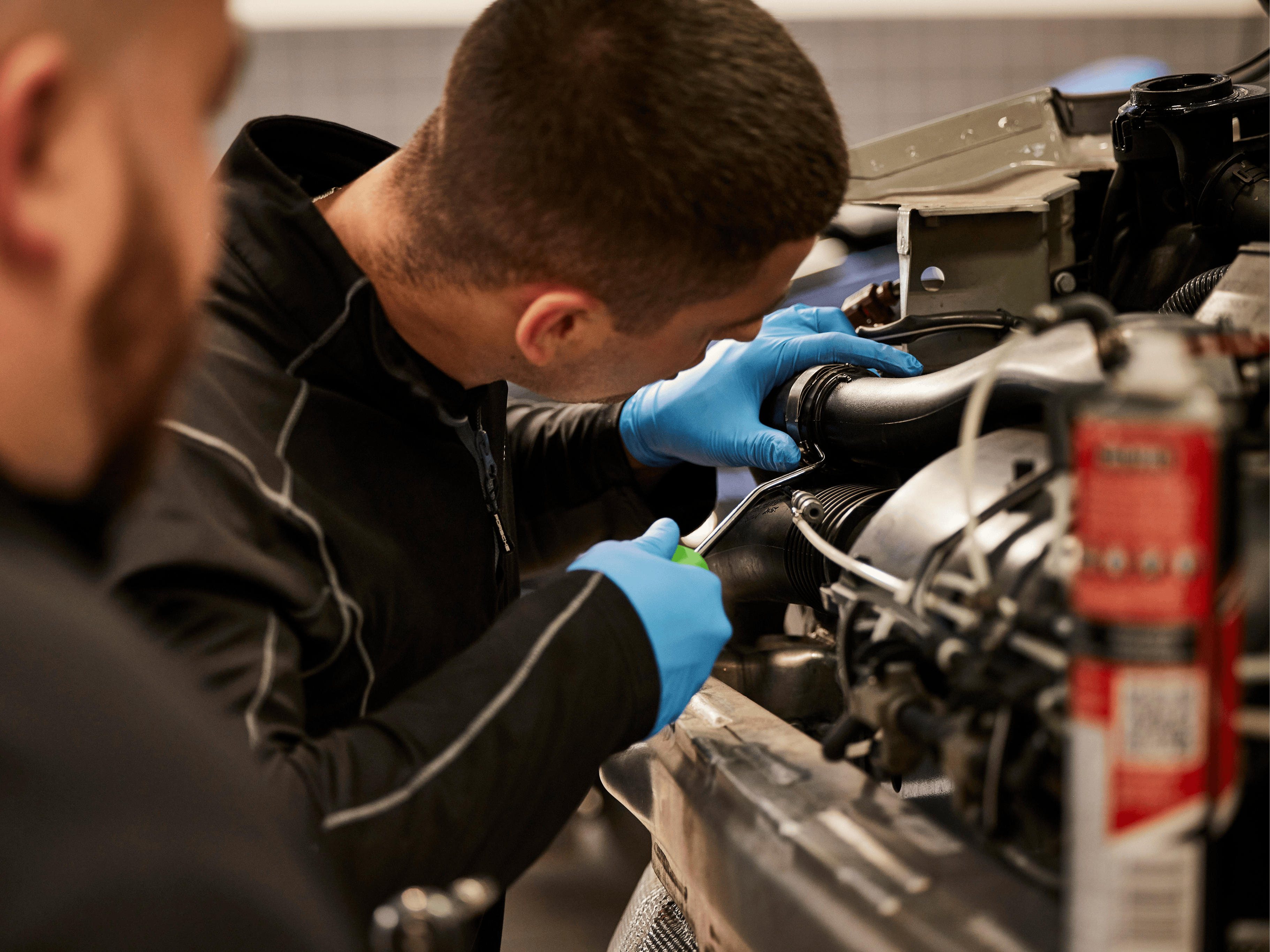 Porsche technician with blue gloves works closely on complex vehicle engine in workshop.