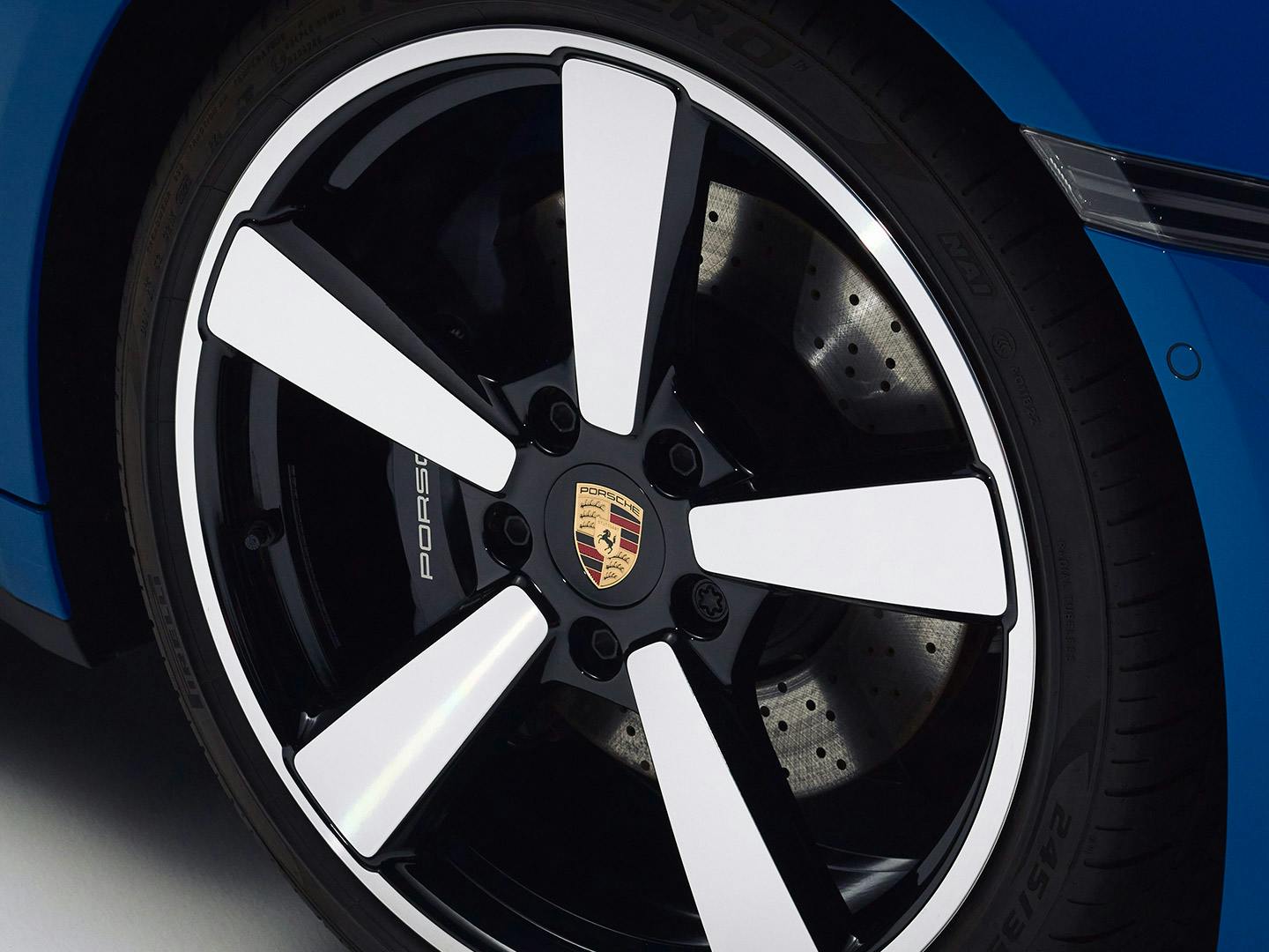 Close-up image of a glossy blue Porsche vehicle's wheel featuring a distinctive black and white five-spoke alloy rim with the Porsche crest in the center.